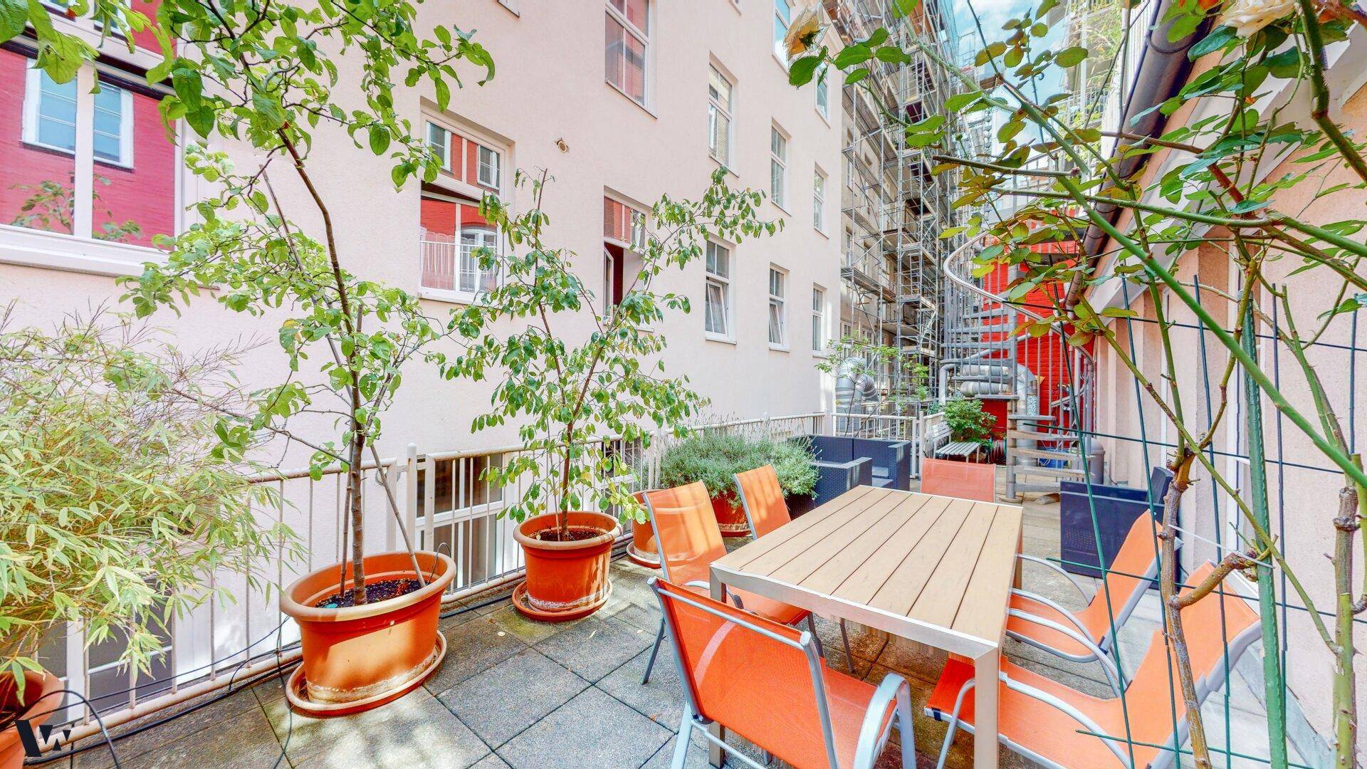 Gemütliche Terrasse mit Esstisch, Stühlen und vielen Topfpflanzen, umgeben von Gebäuden.
