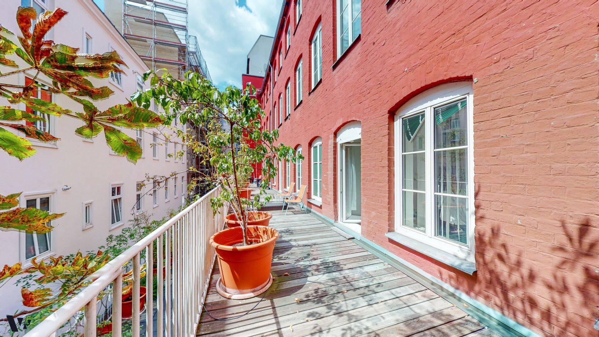 Breiter Balkon mit Holzdeck und Topfpflanzen entlang der roten Backsteinfassade des Gebäudes.