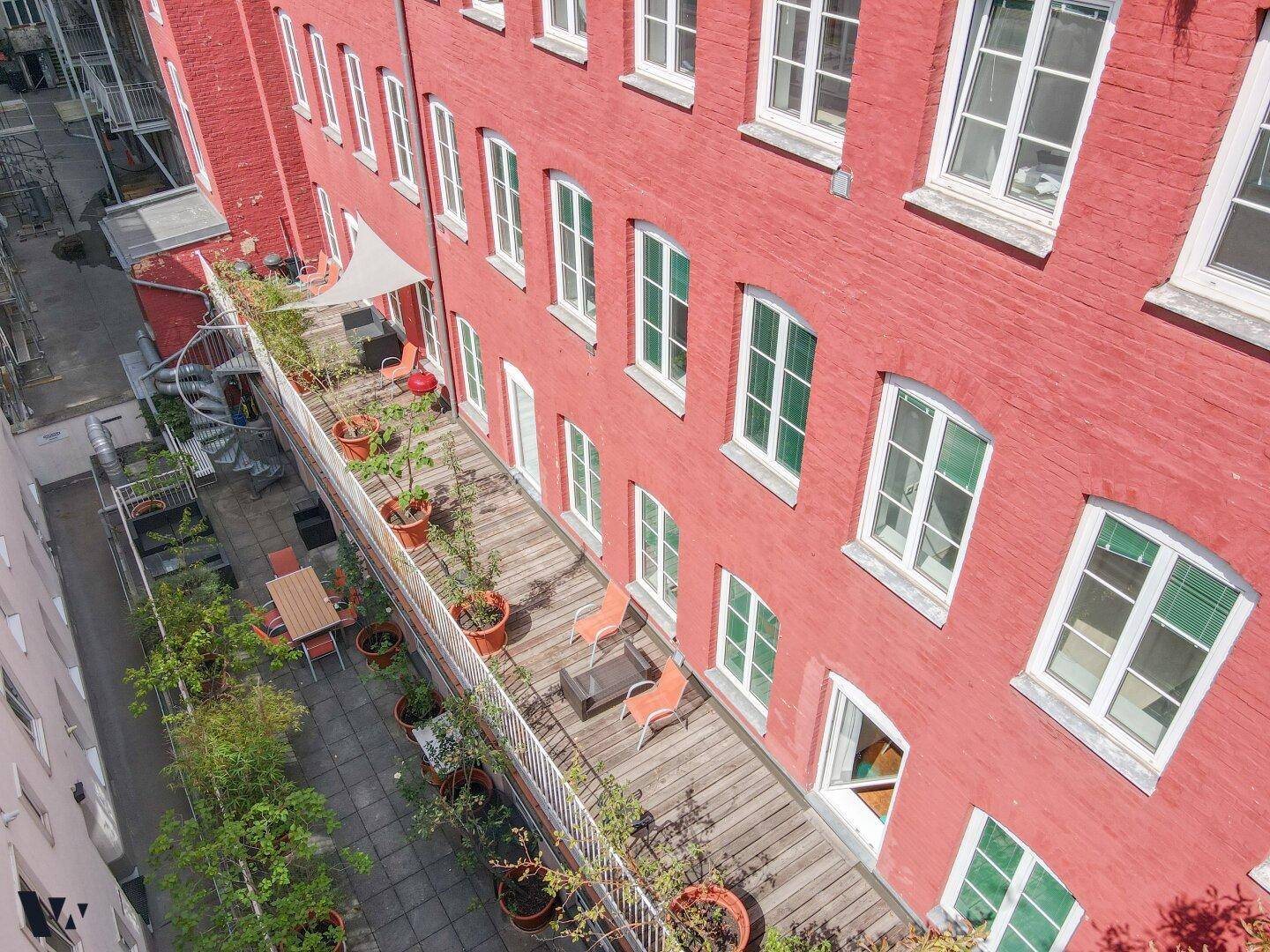 Lange Terrasse mit Holzboden, Pflanzen und Sitzgelegenheiten entlang einer markanten roten Backsteinfassade.