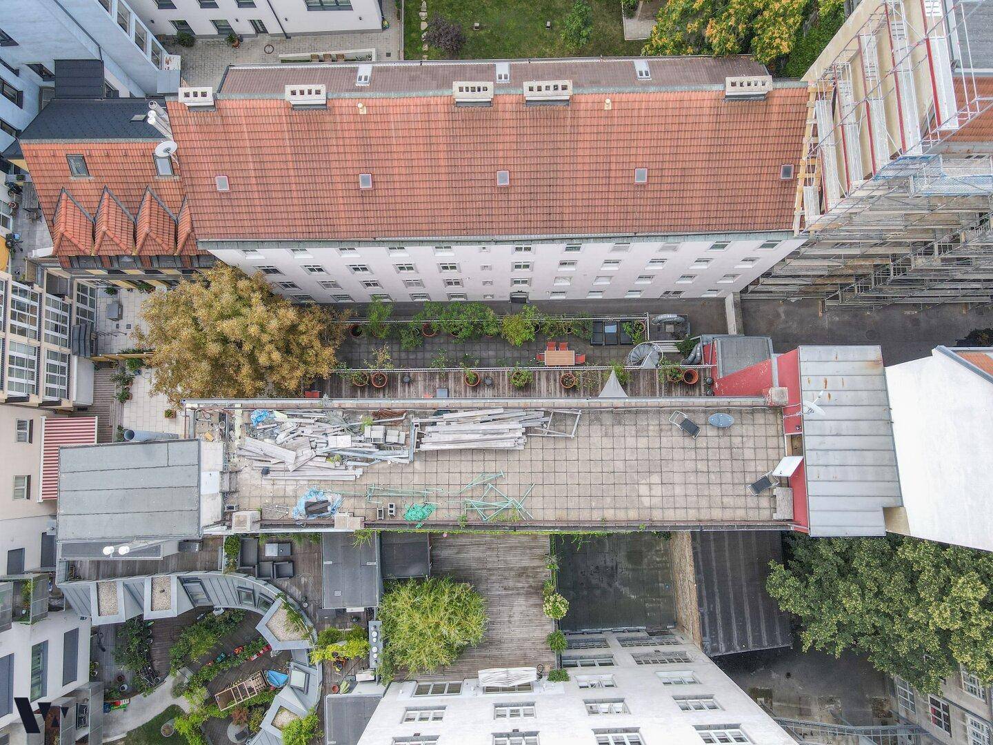Luftaufnahme von Gebäuden mit roten Dächern, grünen Innenhöfen und einer Dachterrasse mit Baumaterialien.