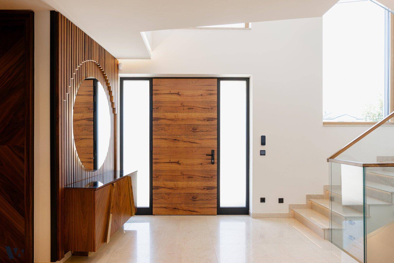 Heller Eingangsbereich mit Holztür, modernem Spiegel und Treppe mit Glasgeländer.