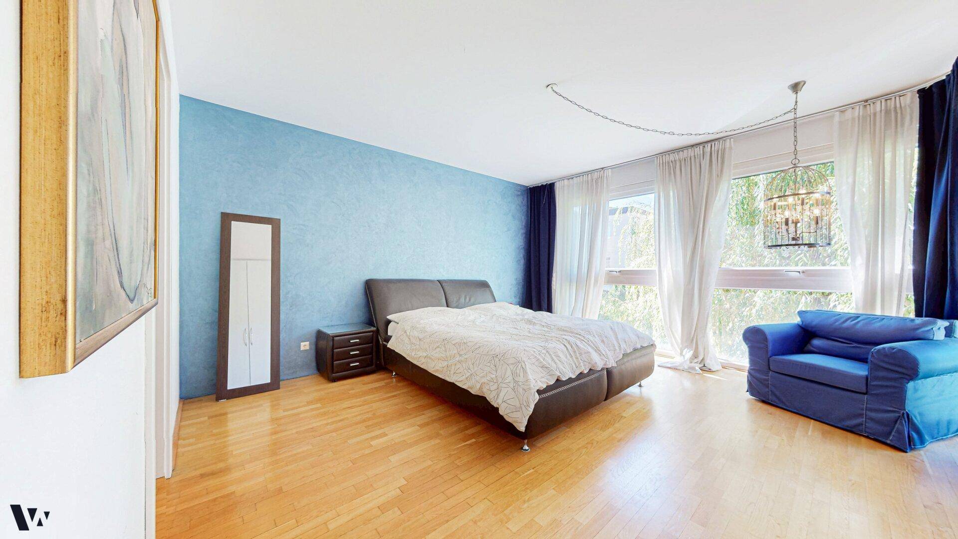 Schlafzimmer mit blauer Akzentwand, Holzboden und großen Fenstern mit viel Tageslicht.
