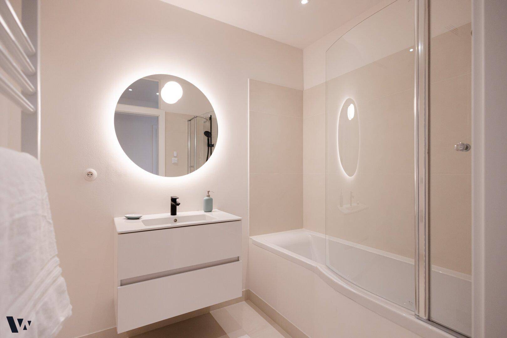 Modernes Badezimmer mit beleuchtetem Spiegel, Waschtisch und Badewanne mit Duschabtrennung.