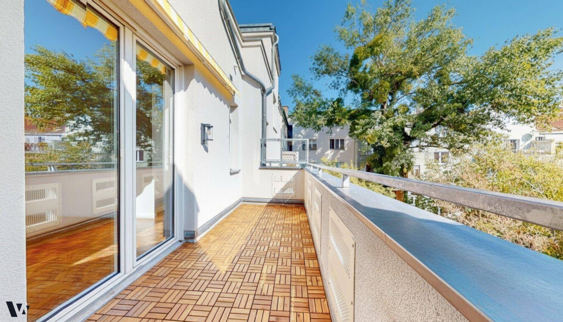 Heller Balkon mit Holzdielen und Schiebetür zum Innenbereich, umgeben von Bäumen.