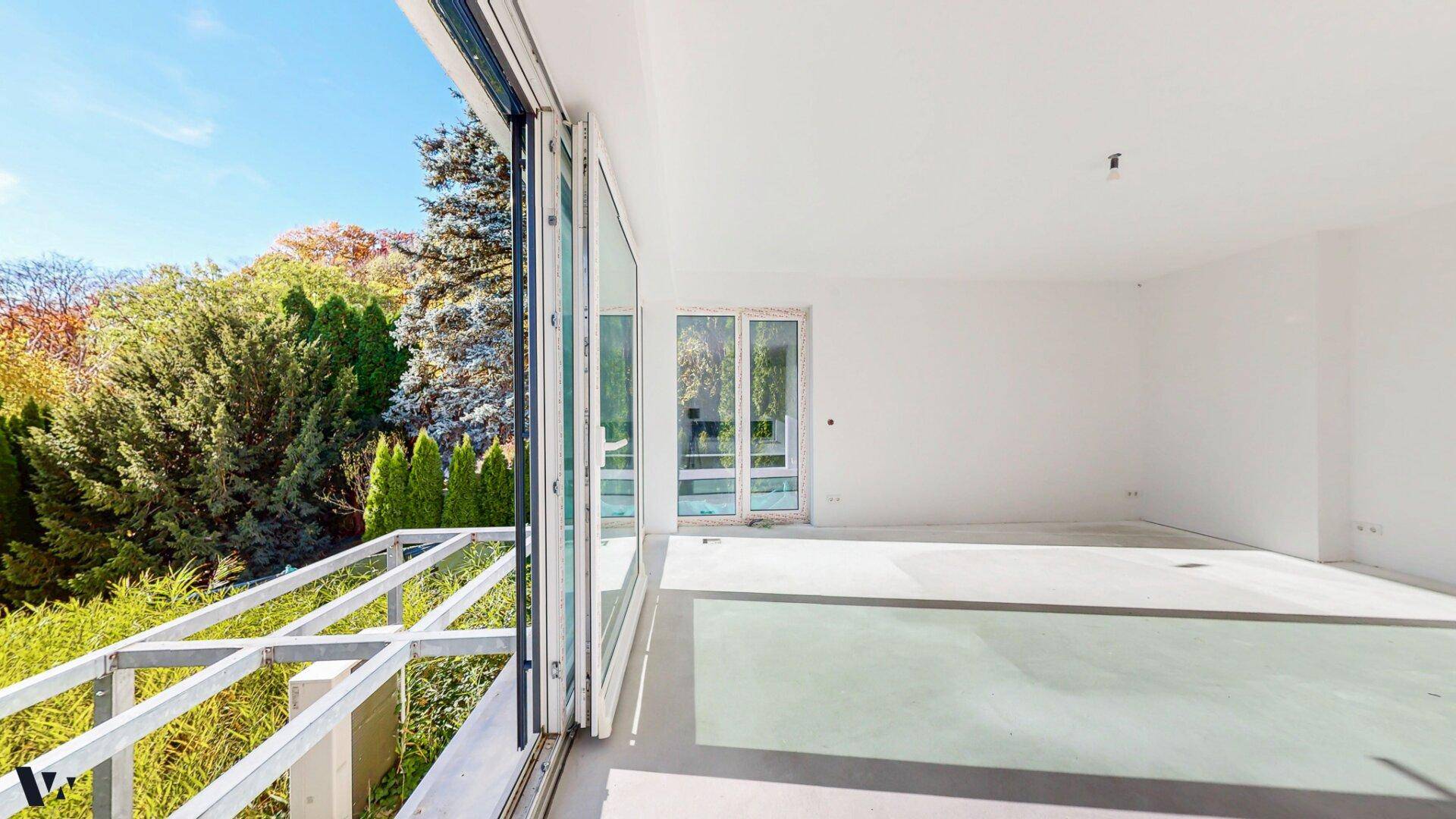 Heller Innenraum mit Schiebetüren, die zu einem Balkon oder einer Terrasse führen, umgeben von Grün.