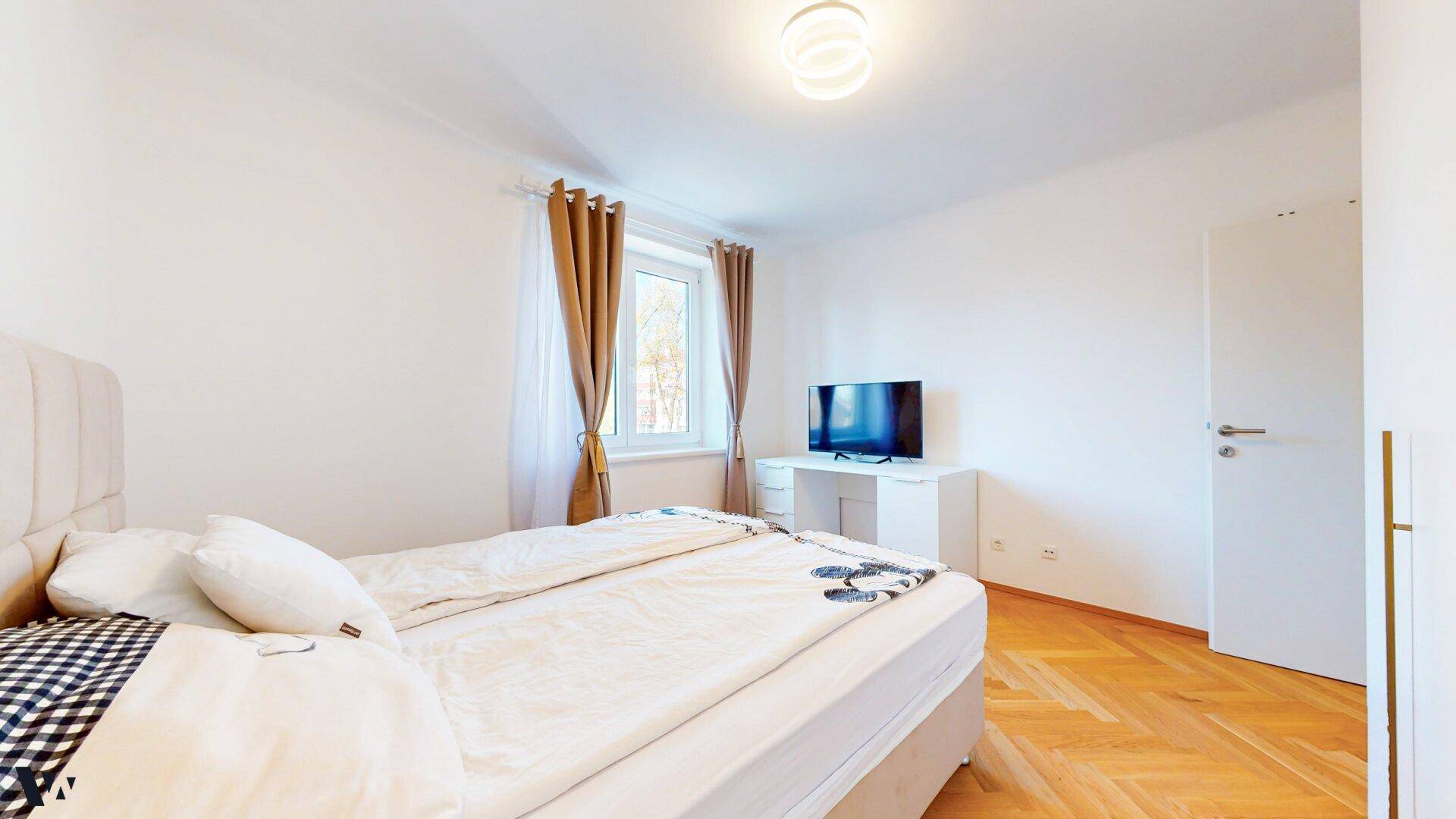 Schlafzimmer mit Parkettboden, Fenster und einem Fernseher auf einem kleinen Schreibtisch.