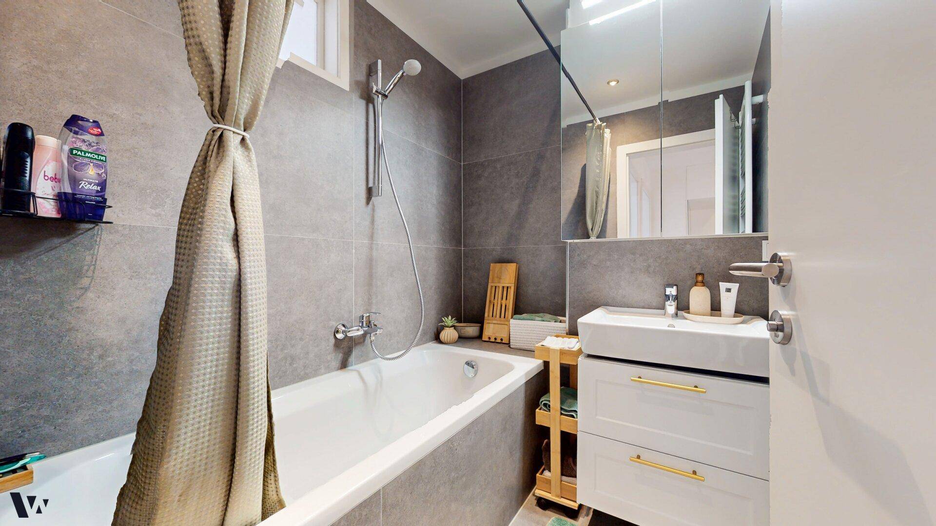 Modernes Badezimmer mit Badewanne, Dusche und grauen Fliesen an Wänden und Boden.
