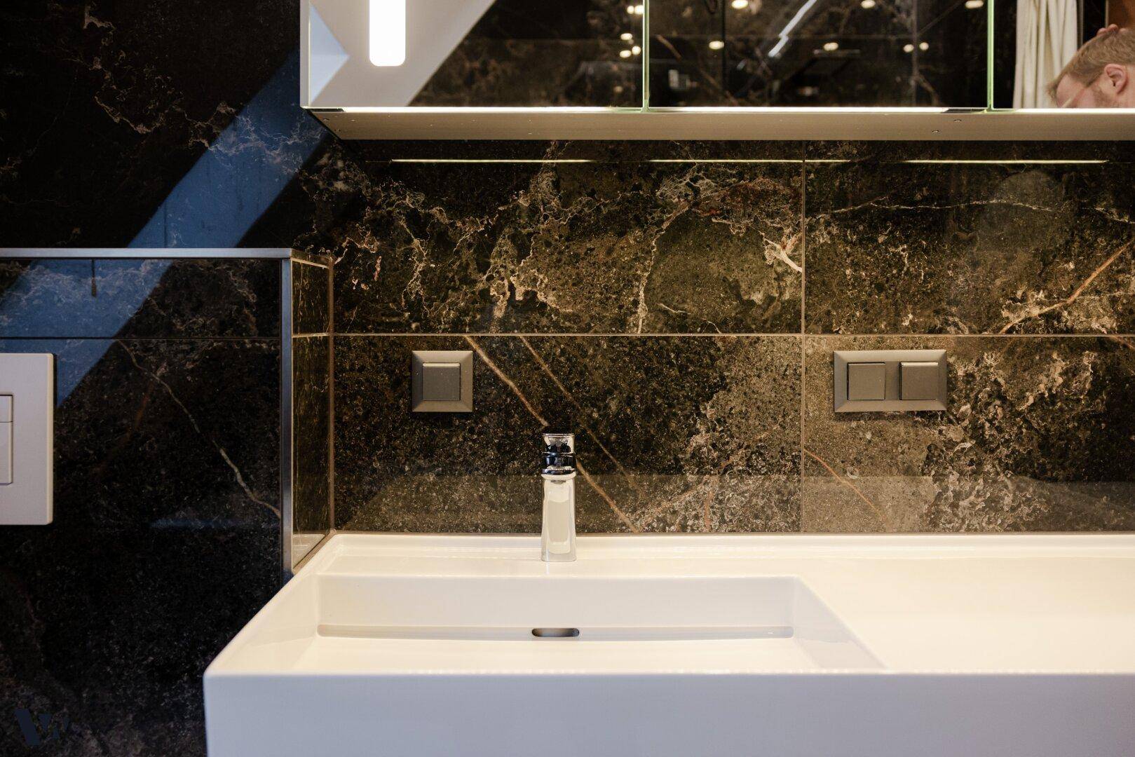 Modernes Badezimmer mit eleganten schwarzen Marmorfliesen, einem weißen Waschbecken und beleuchtetem Spiegelschrank.
