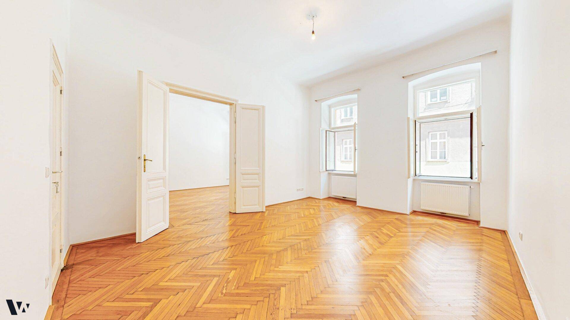 Weitläufiges, helles Zimmer mit Fischgrätparkett und hohen Flügeltüren.