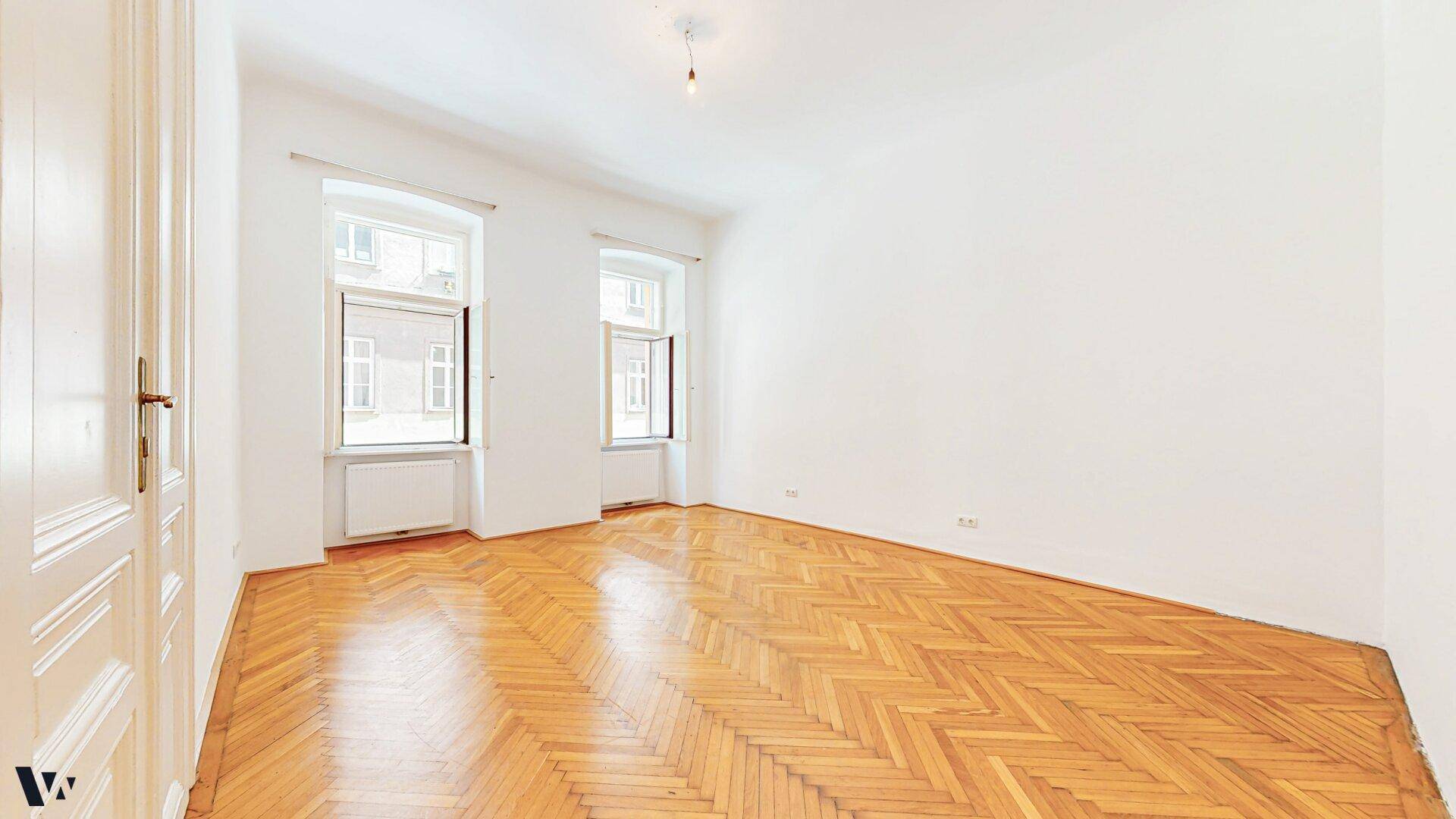 Helles Zimmer mit Fischgrätparkett und zwei hohen Fenstern, ideal für Wohn- oder Schlafbereich.