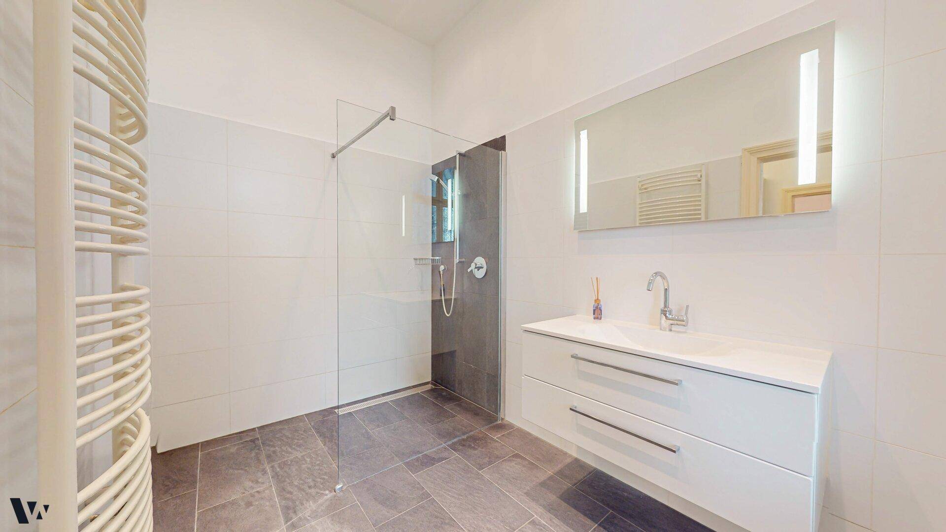 Zeitgemäßes Badezimmer mit großer Dusche, modernem Waschtisch und beleuchtetem Spiegel.
