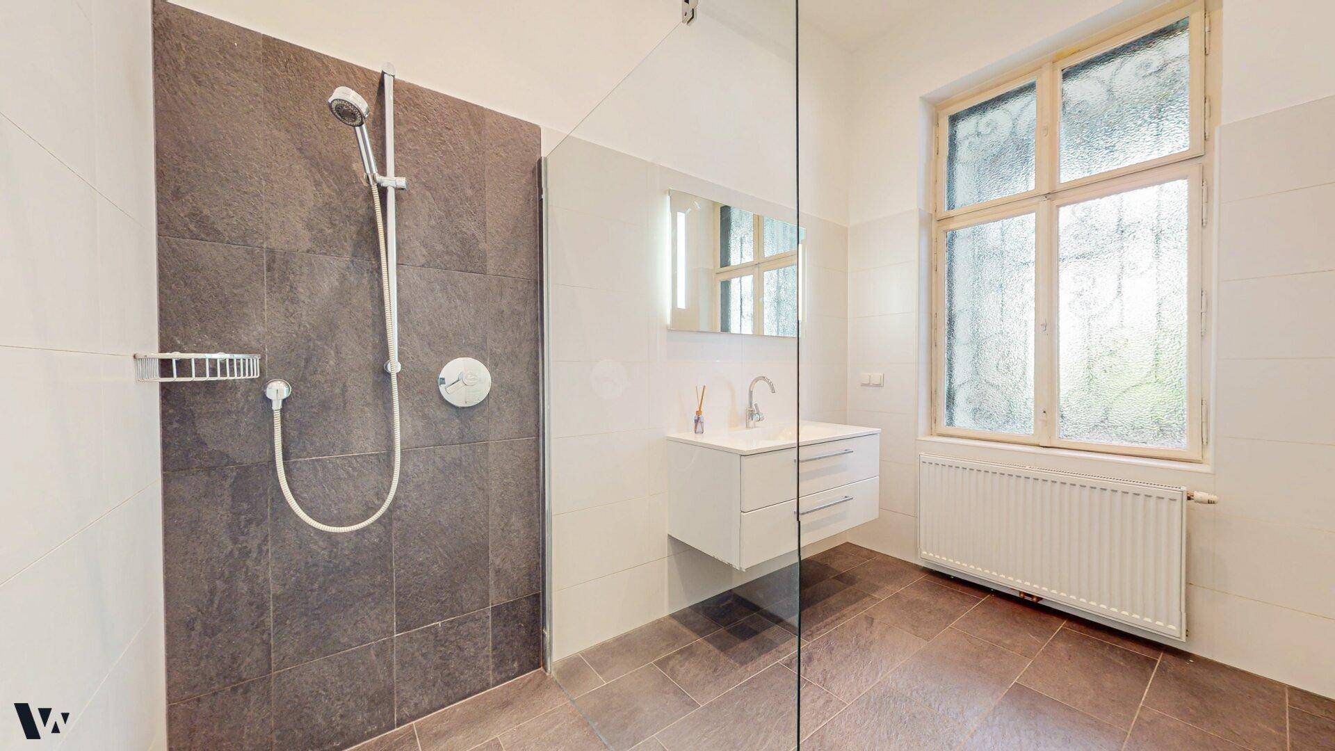 Helles Badezimmer mit großer Dusche, Waschtisch und Fenster für natürliche Belüftung.