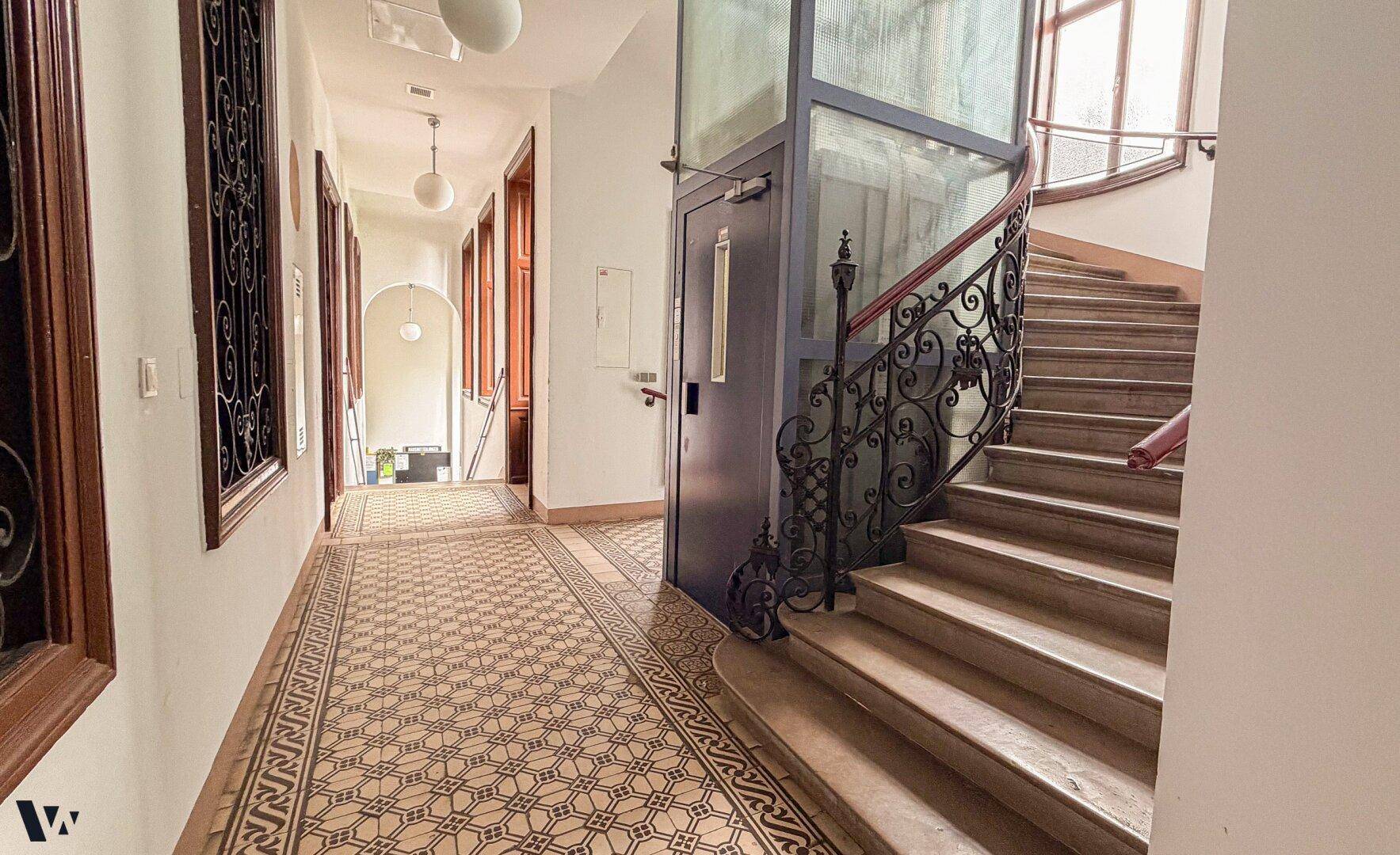 Historisches Treppenhaus mit kunstvollem Geländer, Aufzug und gemustertem Fliesenboden.
