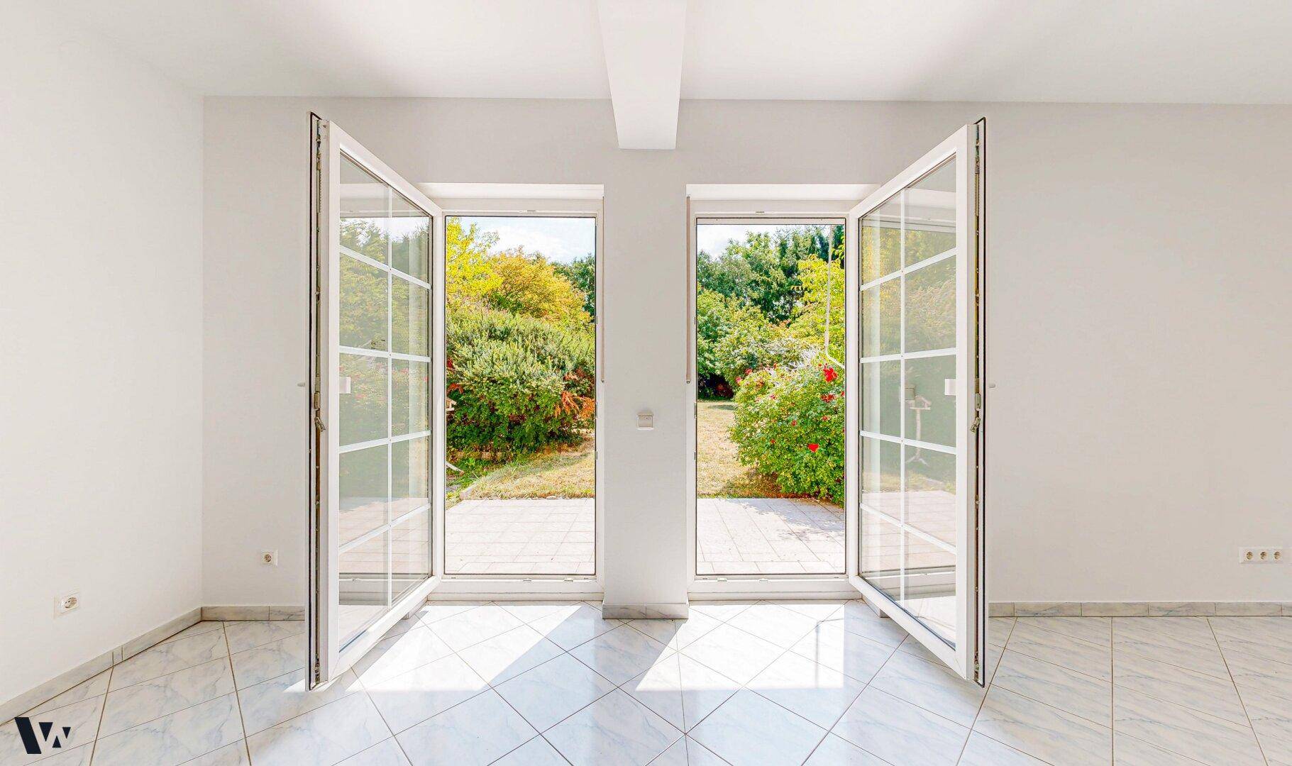 Heller Raum mit großen, geöffneten Fenstertüren, die direkten Zugang zum grünen Garten bieten.