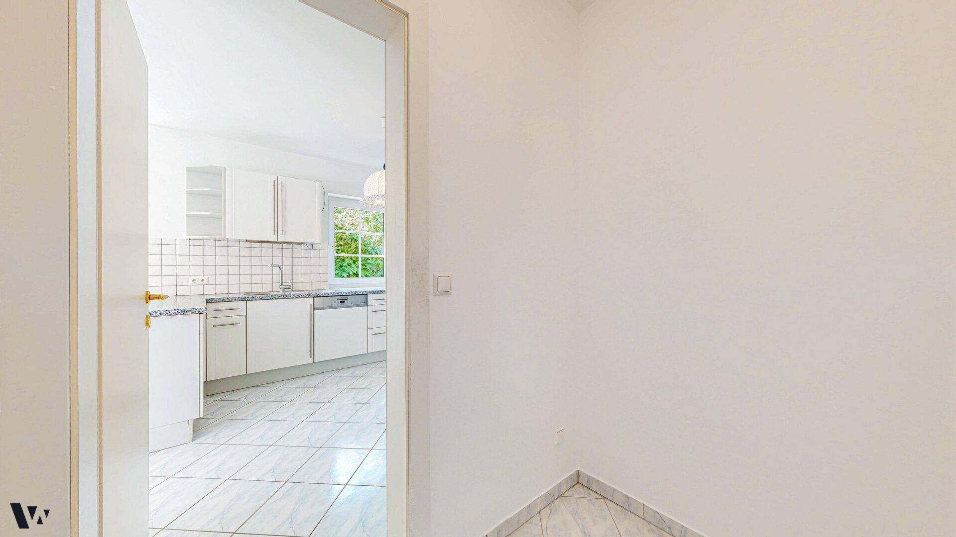 Blick vom Flur in die helle, modern ausgestattete Küche mit weißen Schränken und Fliesenboden.