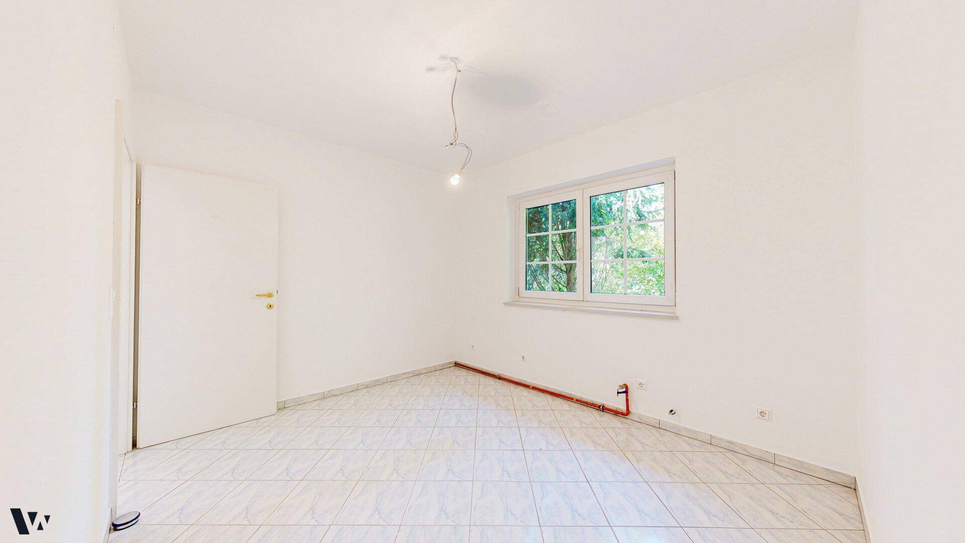 Helles Zimmer mit weißen Wänden, Fliesenboden und einem Fenster mit Blick ins Grüne.