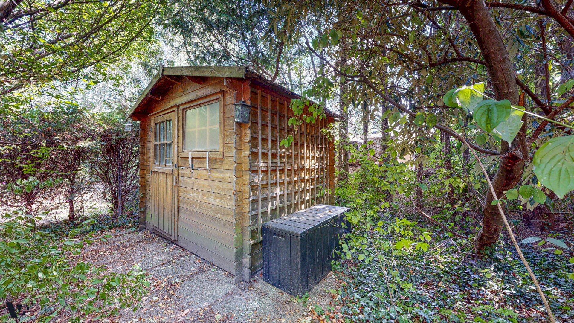 Rustikaler Holzschuppen im Garten, umgeben von Bäumen und Sträuchern, ideal für Gartengeräte.