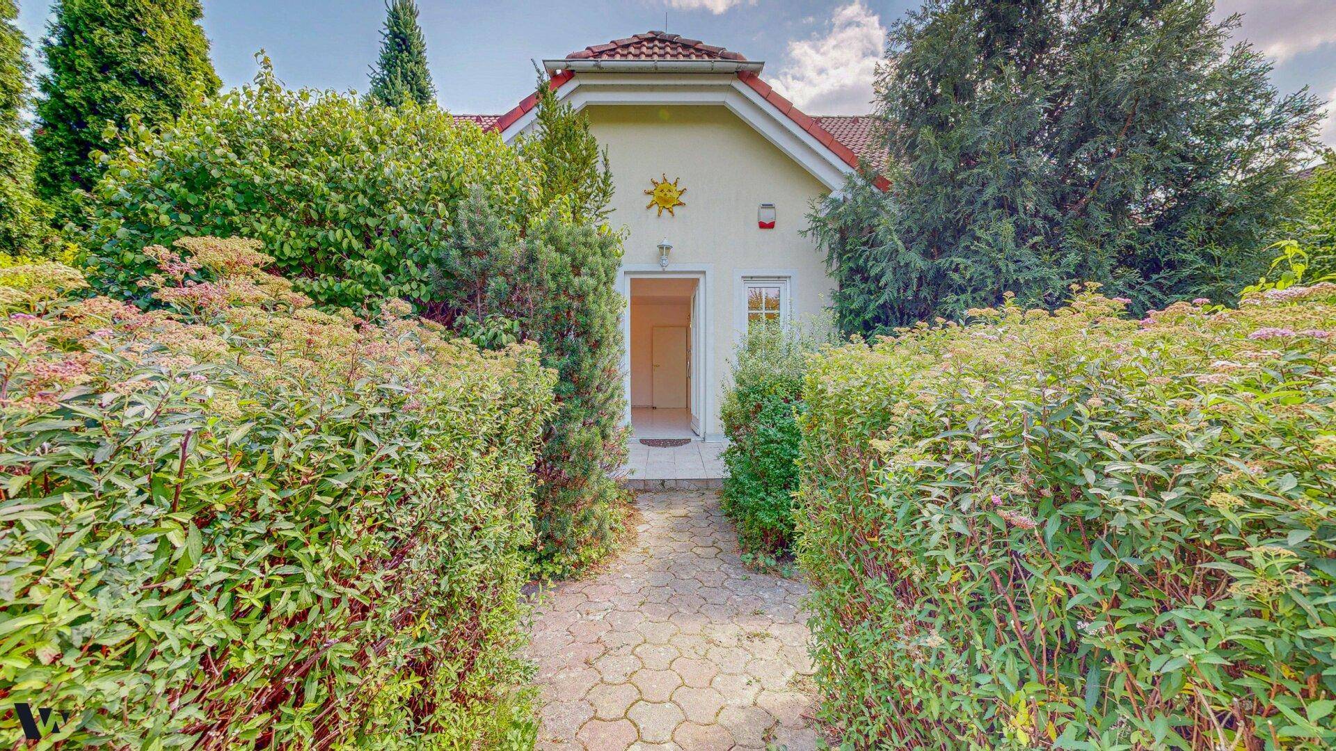 Eingangsbereich des Hauses mit einem gepflasterten Weg, umgeben von dichten grünen Hecken.
