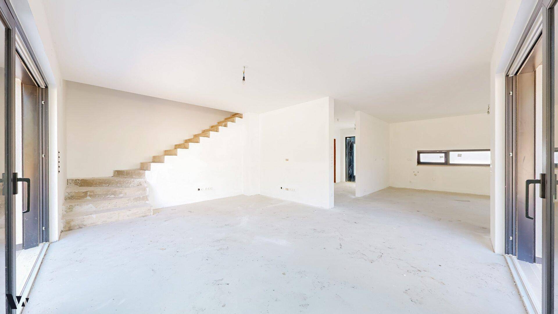 Geräumiger, unmöblierter Wohnbereich im Rohbau mit Treppe und großen Terrassentüren.
