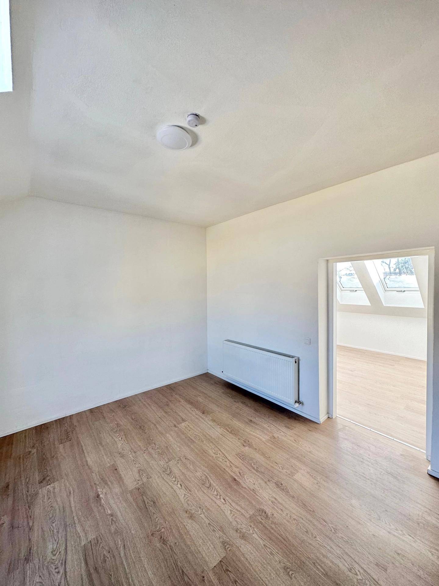 Helles Zimmer mit Laminatboden und Heizkörper, offen zu einem weiteren Raum mit Dachschrägen.