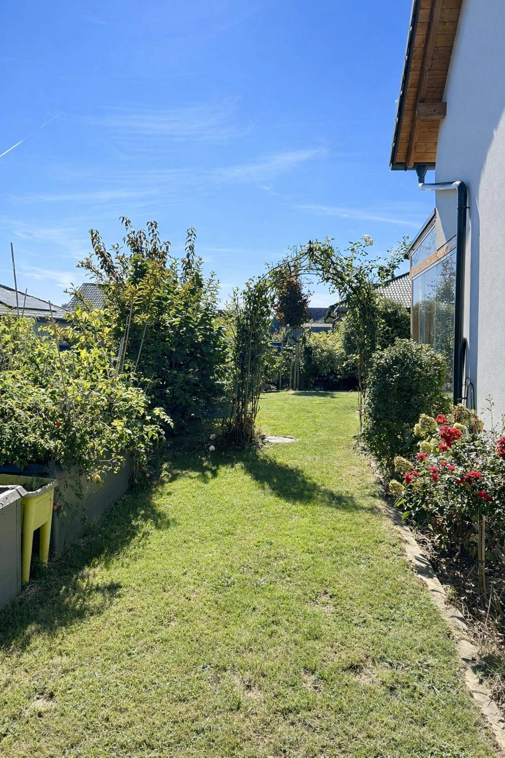 Langer Gartenweg mit Rasenflächen und einem Rosenbogen, umgeben von Sträuchern.