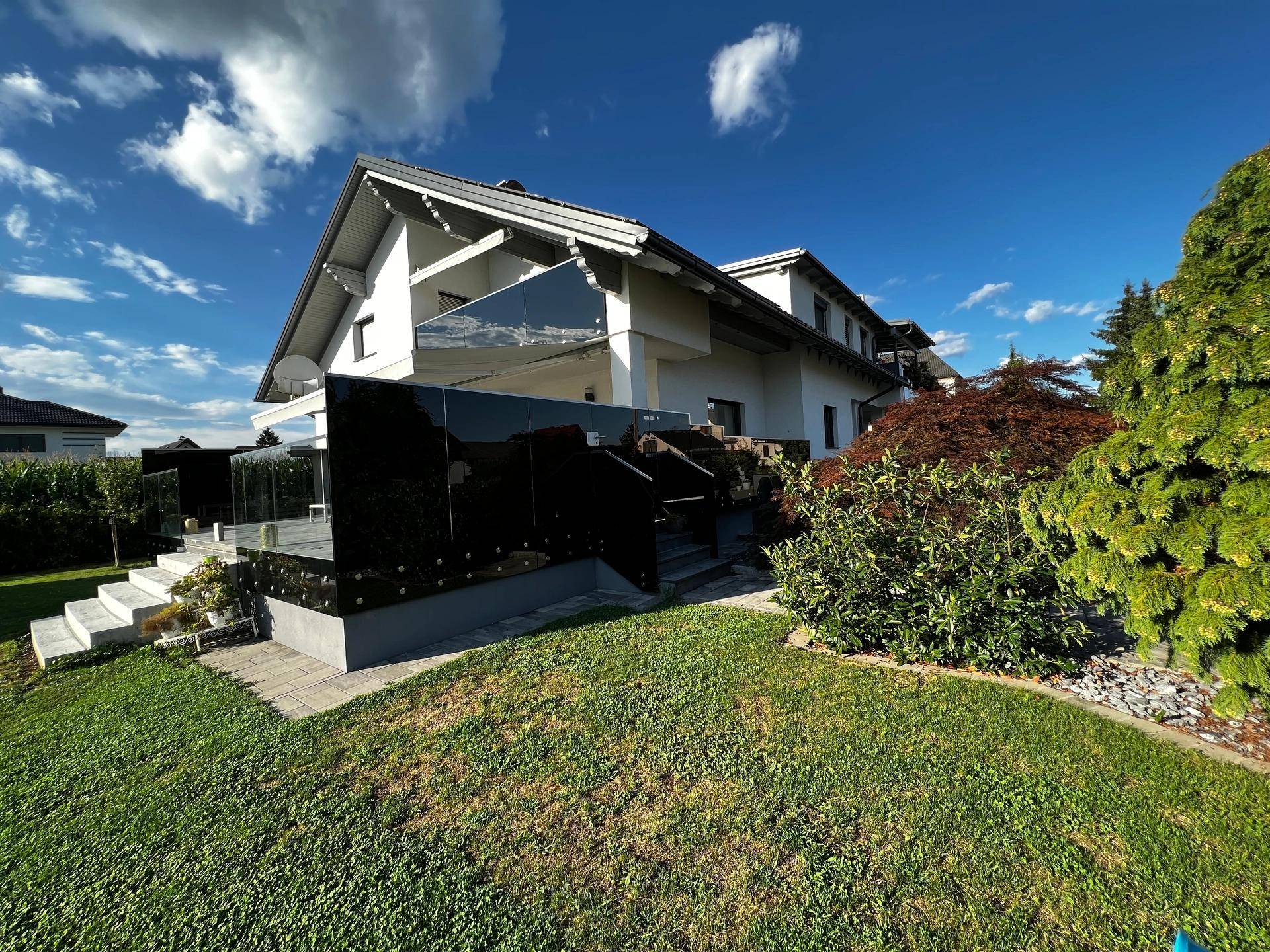 Seitlicher Blick auf das Haus mit gepflegtem Garten, Terrasse und moderner Glasbrüstung.