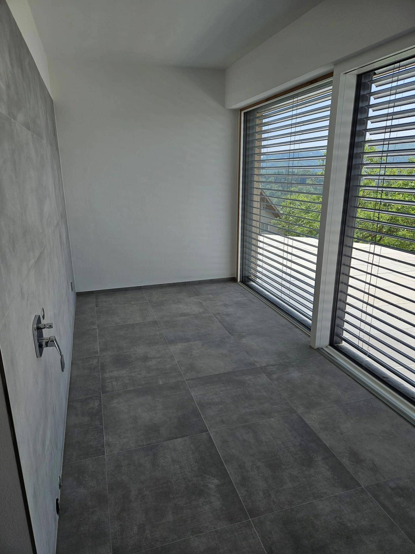 Modernes Badezimmer mit grauen Fliesen, großer Fensterfront und Blick ins Grüne.