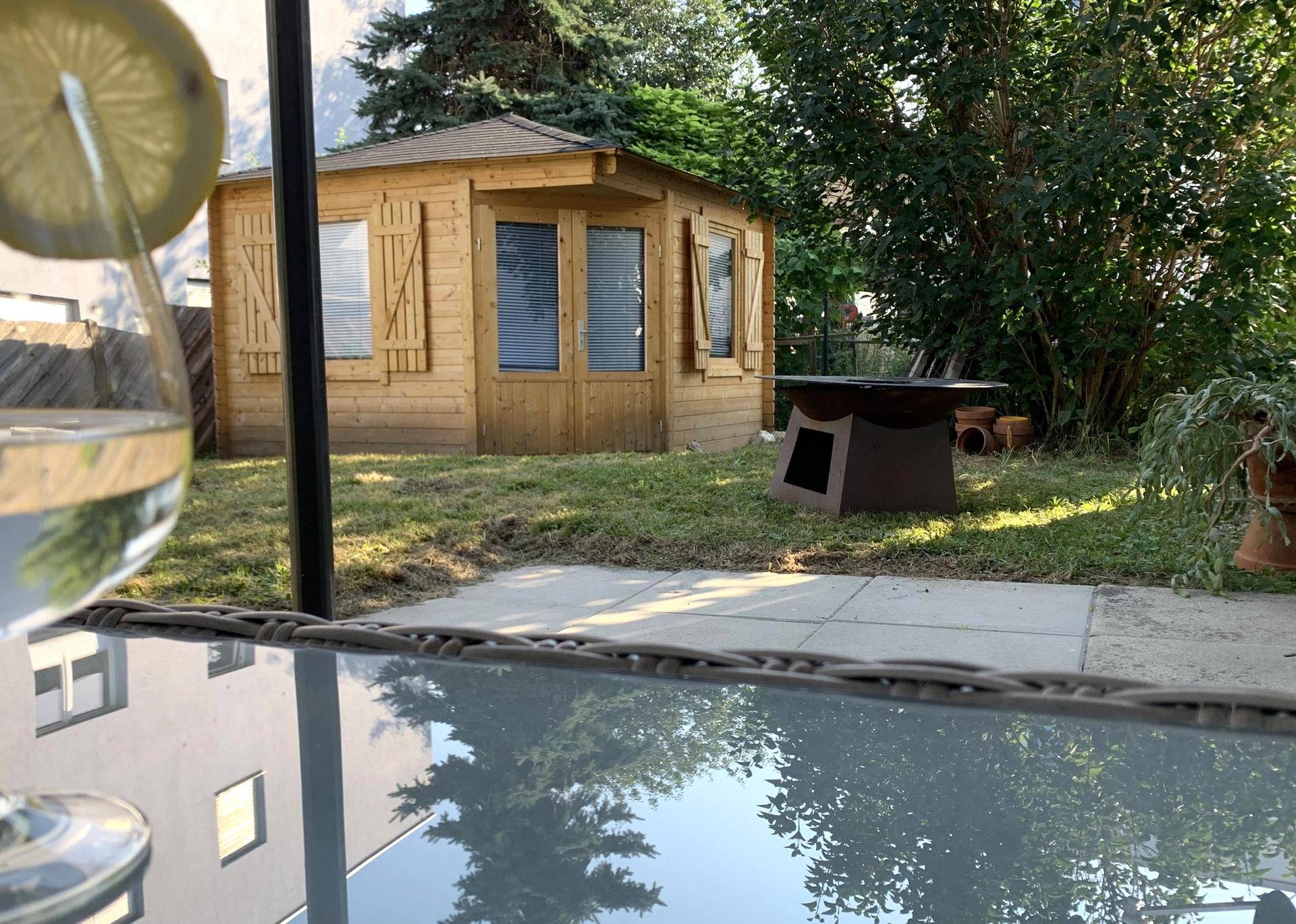 Blick von der Terrasse auf den Garten mit einem Holzgartenhaus und einer modernen Feuerstelle.