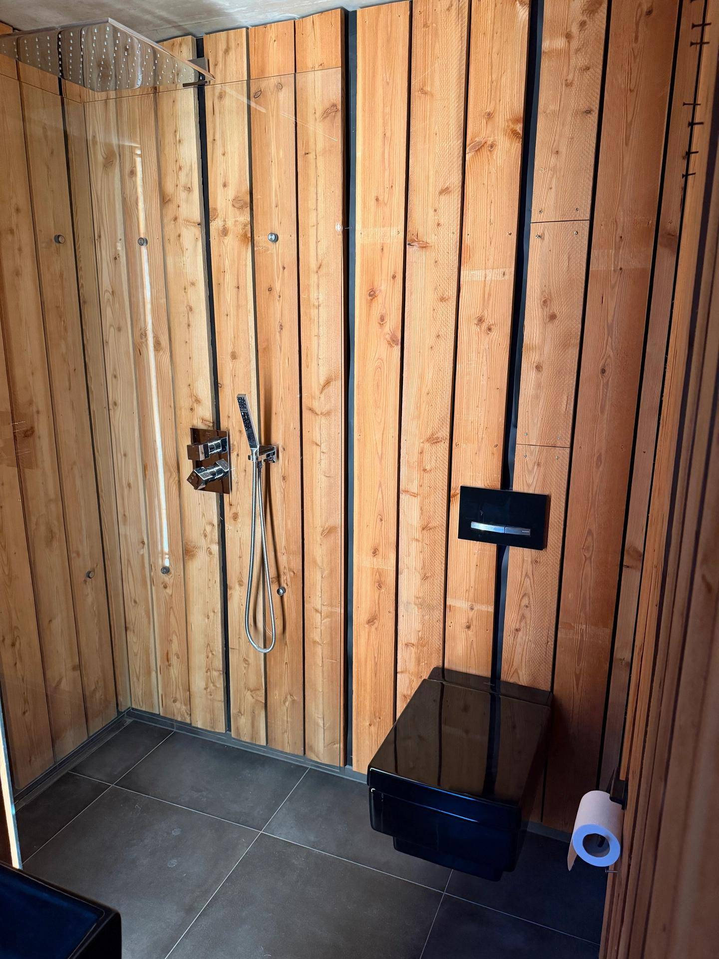 Elegantes Badezimmer mit Holzwänden, dunklen Fliesen, einer begehbaren Regendusche und Wand-WC.