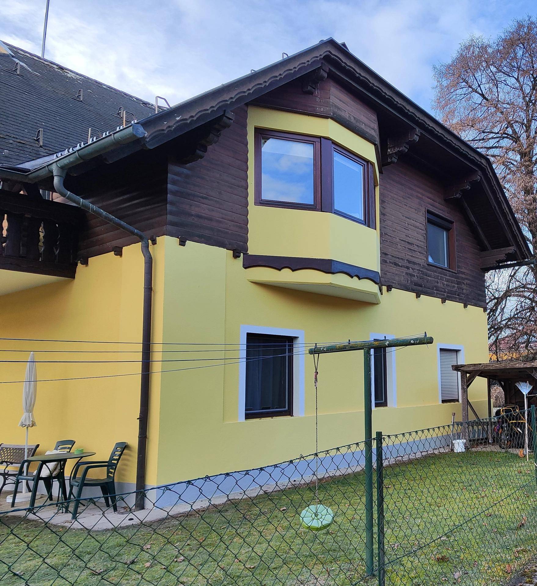 Seitenansicht des gelben Gebäudes mit Holzelementen, Erker und kleinem Außenbereich mit Tisch.