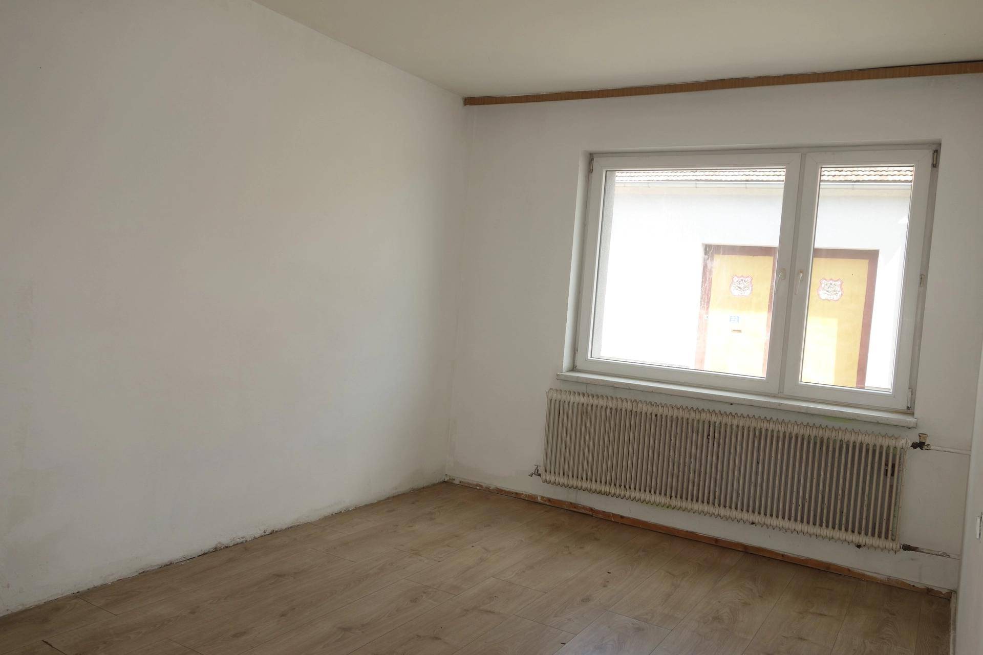 Helles, leeres Zimmer mit weißen Wänden, Laminatboden, Fenster und Heizkörper.