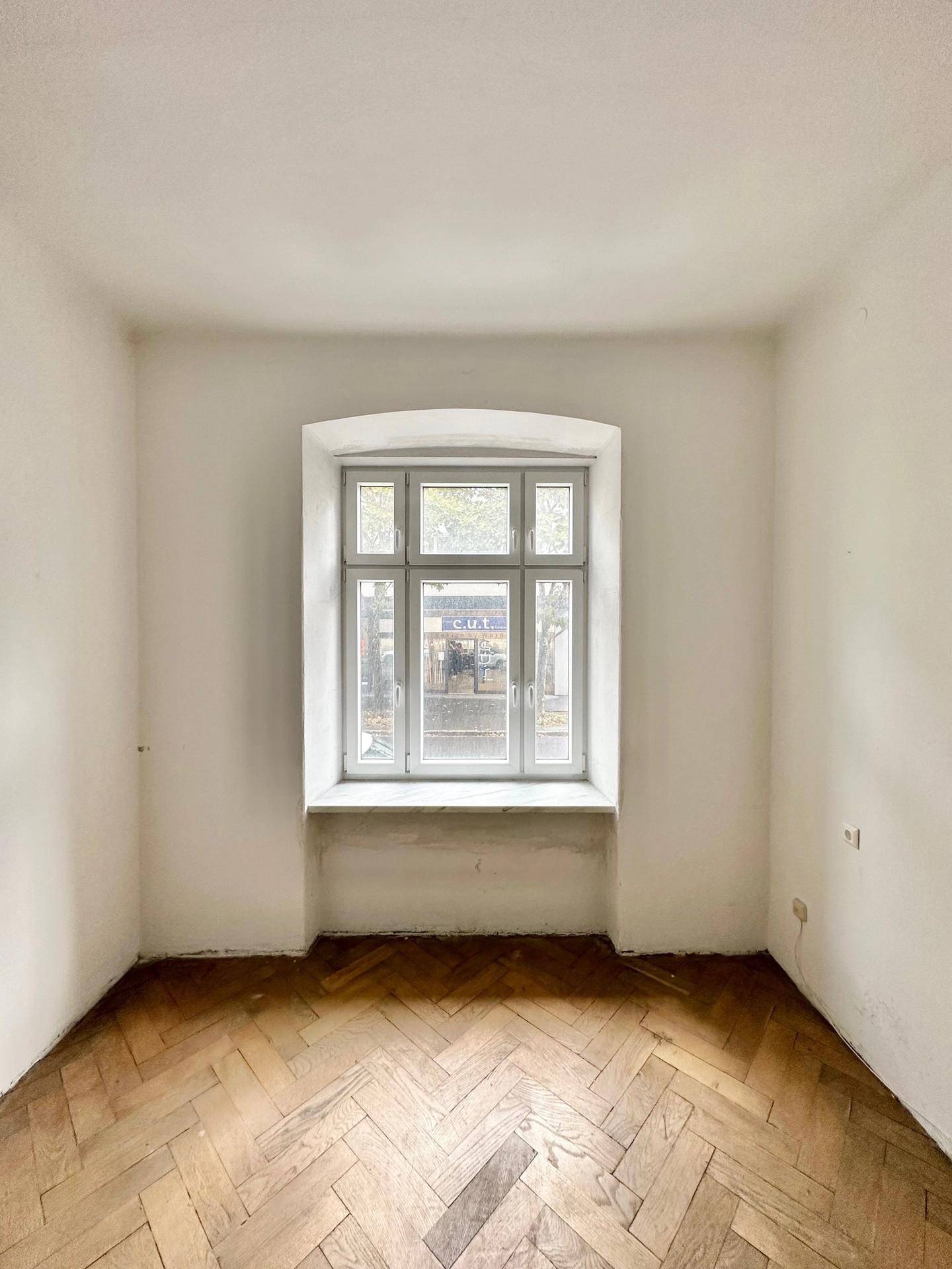 Heller Raum mit Fischgrätparkett und großem Fenster zur Straße.