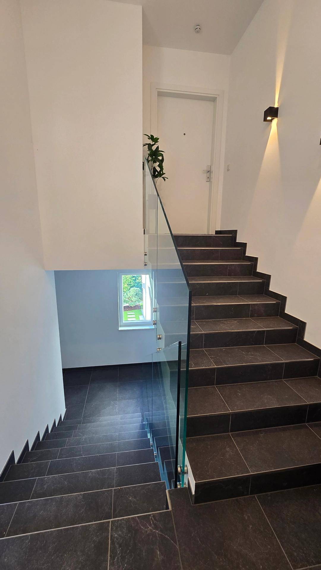 Elegantes Treppenhaus mit dunklen Fliesen und modernem Glasgeländer, gut beleuchtet.