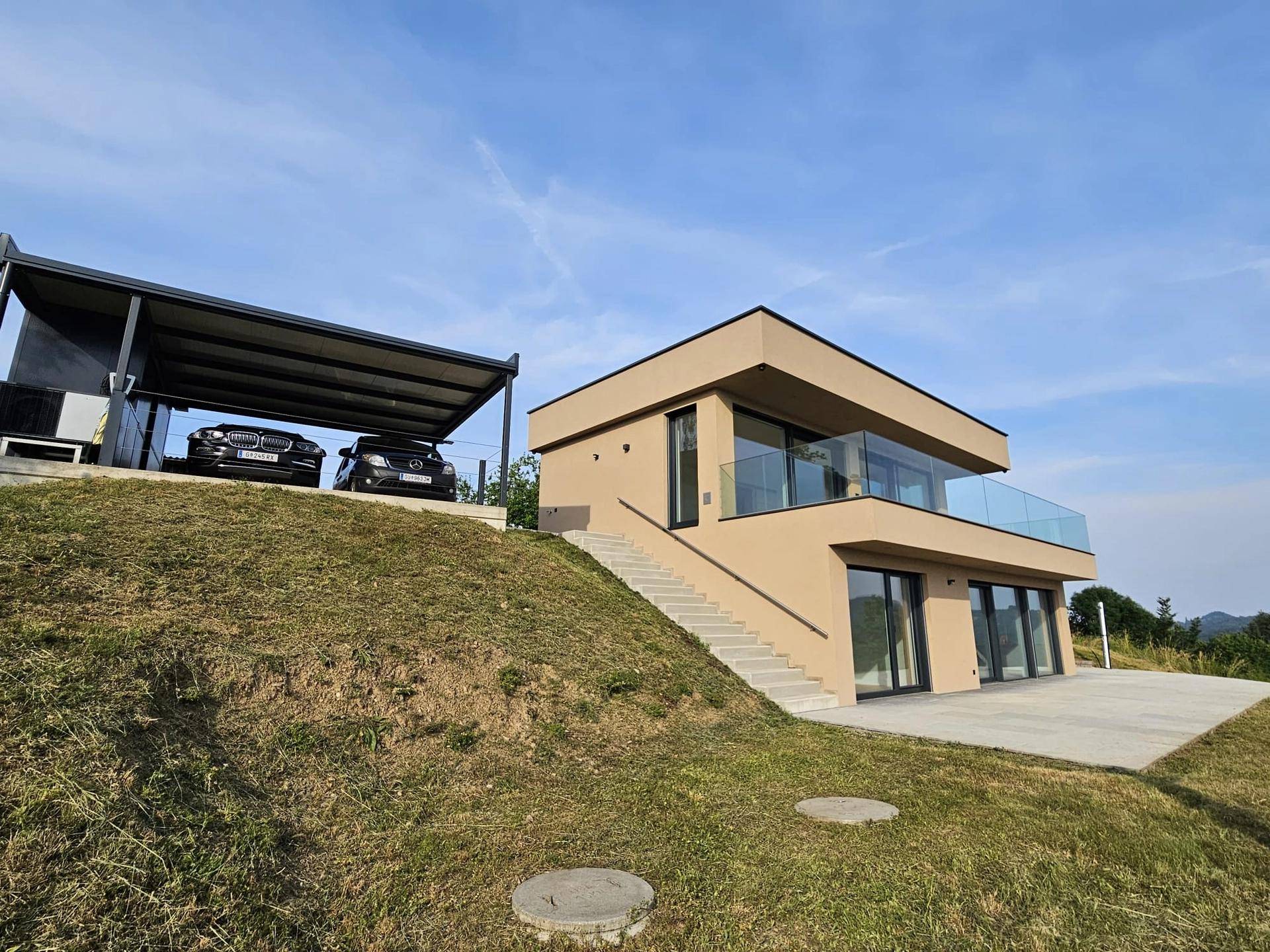 Modernes Haus mit Carport und weitläufiger Grünfläche, eingebettet in eine idyllische Hügellandschaft.