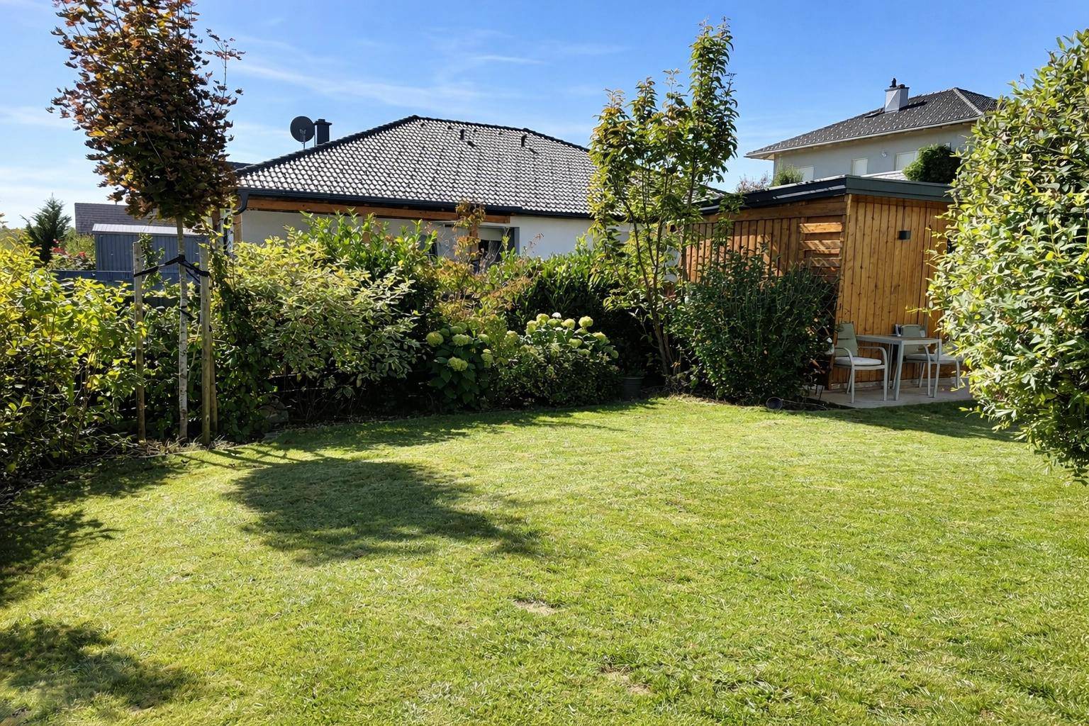 Gepflegter Garten mit Rasenfläche, Sträuchern und einem kleinen Sitzbereich im Hintergrund.