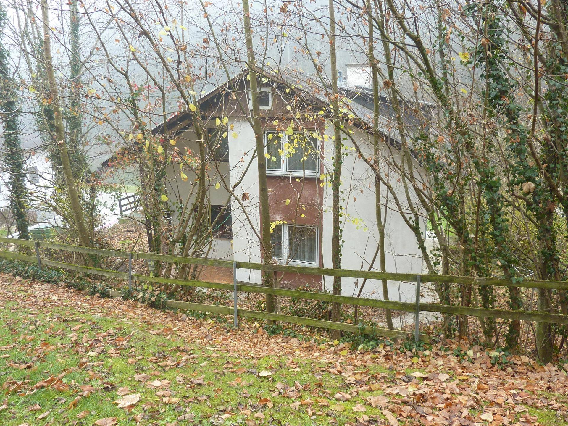 Hausfassade, teilweise verdeckt von kahlen Bäumen und einem Holzzaun im Herbst.