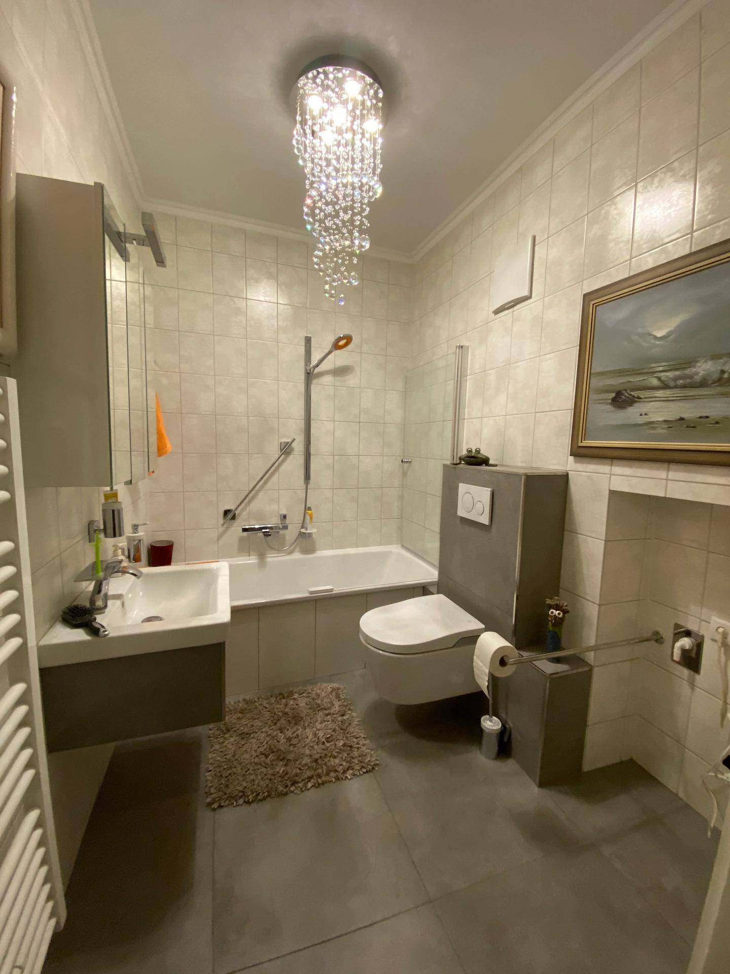 Modernes Badezimmer mit Badewanne, wandhängendem WC, Handtuchheizkörper und elegantem Kronleuchter.