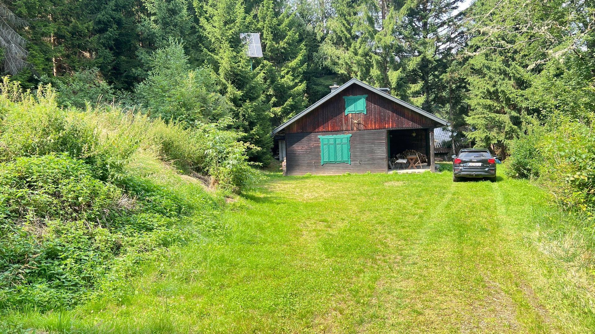 Charmantes Holzhaus mit grünen Fensterläden, umgeben von üppiger Natur und einem geparkten Fahrzeug.