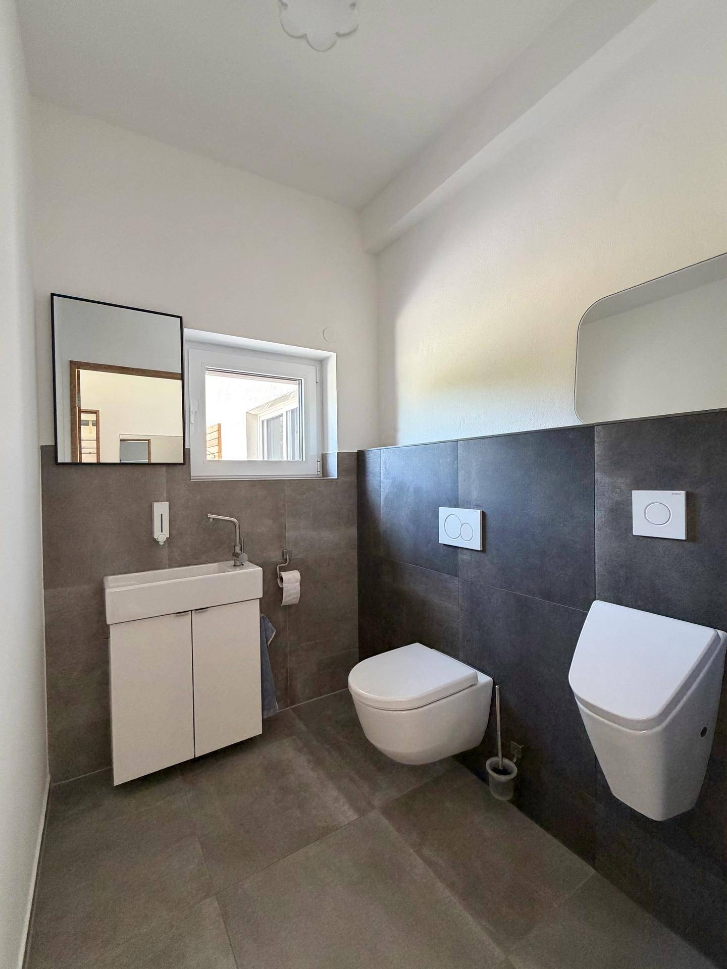 Modernes Gäste-WC mit dunklen Fliesen, wandhängender Toilette, Urinal und kleinem Fenster.