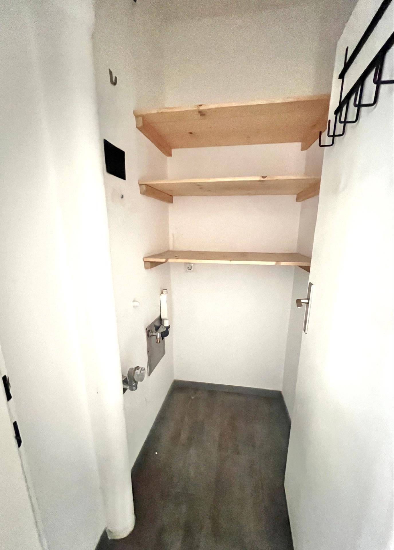 Kleiner Abstellraum mit Holzregalen, Anschlüssen für Waschmaschine und dunklem Boden.