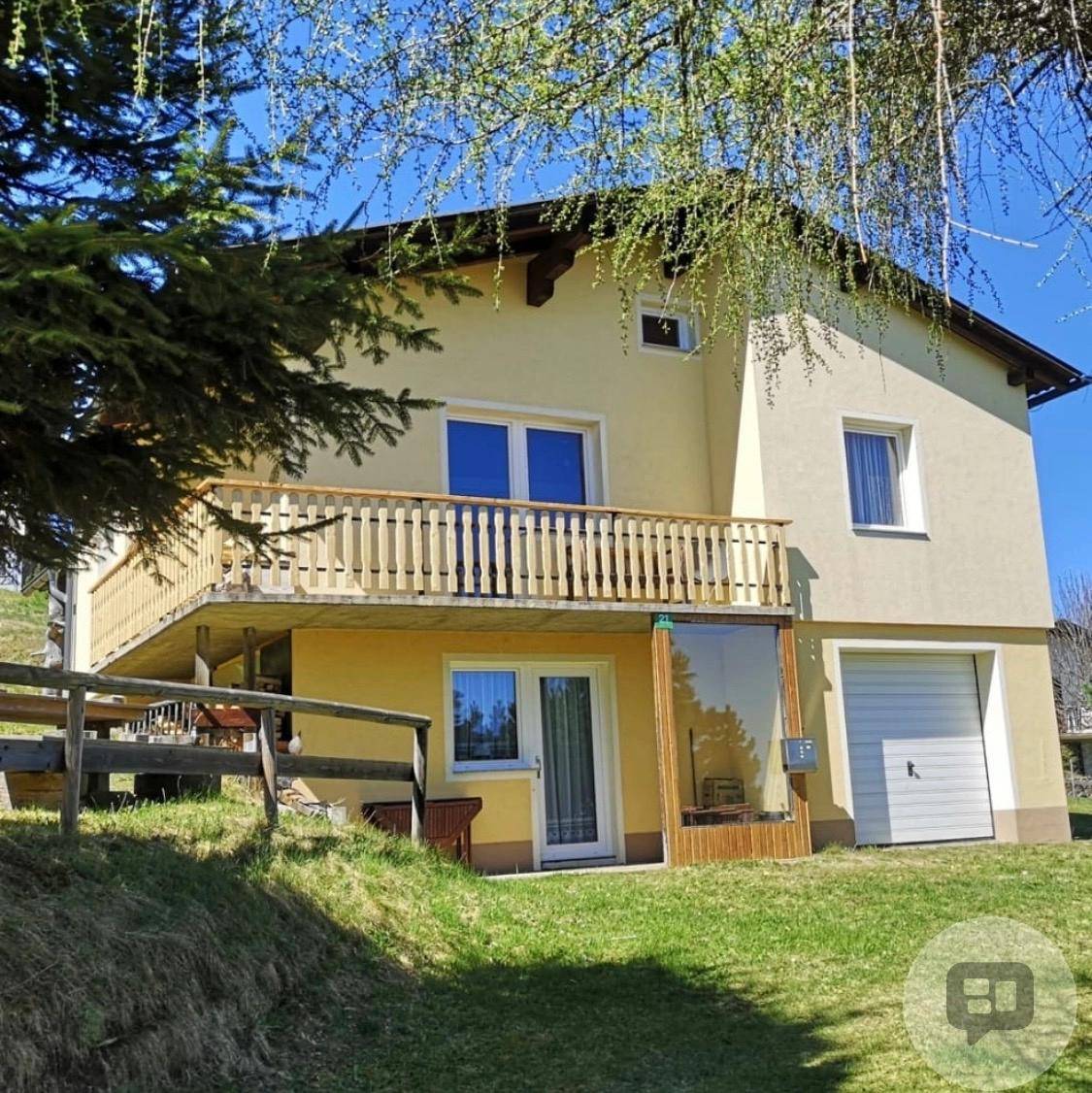 Einfamilienhaus mit Balkon, Garage und grünem Garten in ländlicher Umgebung an einem sonnigen Tag.