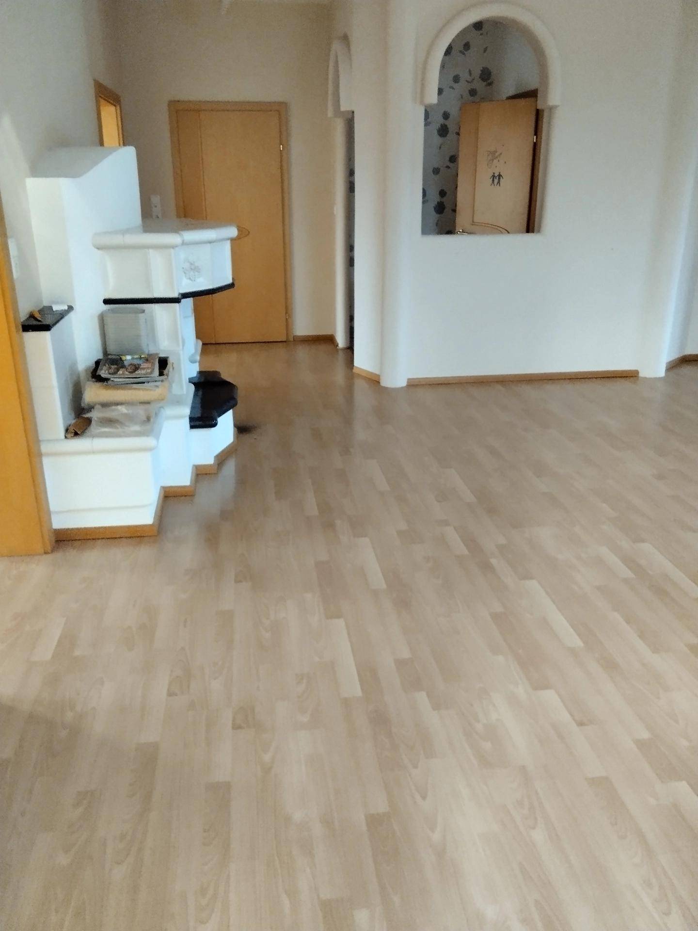 Heller Wohnbereich mit Laminatboden, einem dekorativen Kachelofen und Rundbögen.