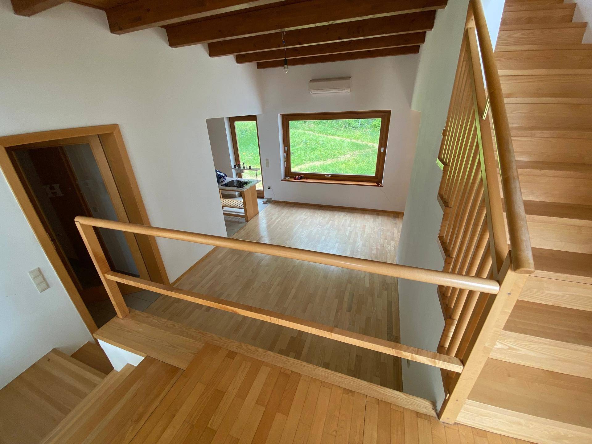 Blick von der oberen Etage über ein Holzgeländer auf den unteren Wohnbereich mit Fenstern.