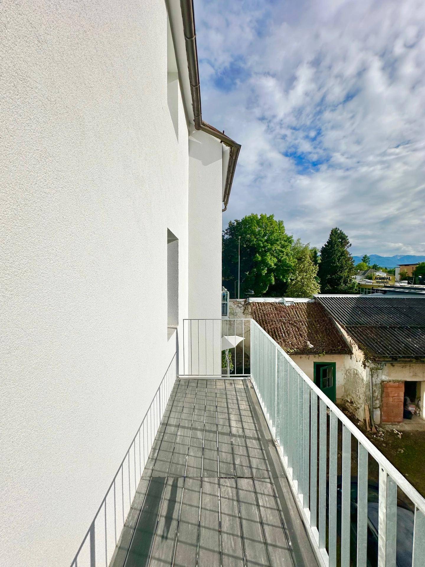 Langer, schmaler Balkon mit weißem Geländer und Blick auf die Umgebung unter bewölktem Himmel.
