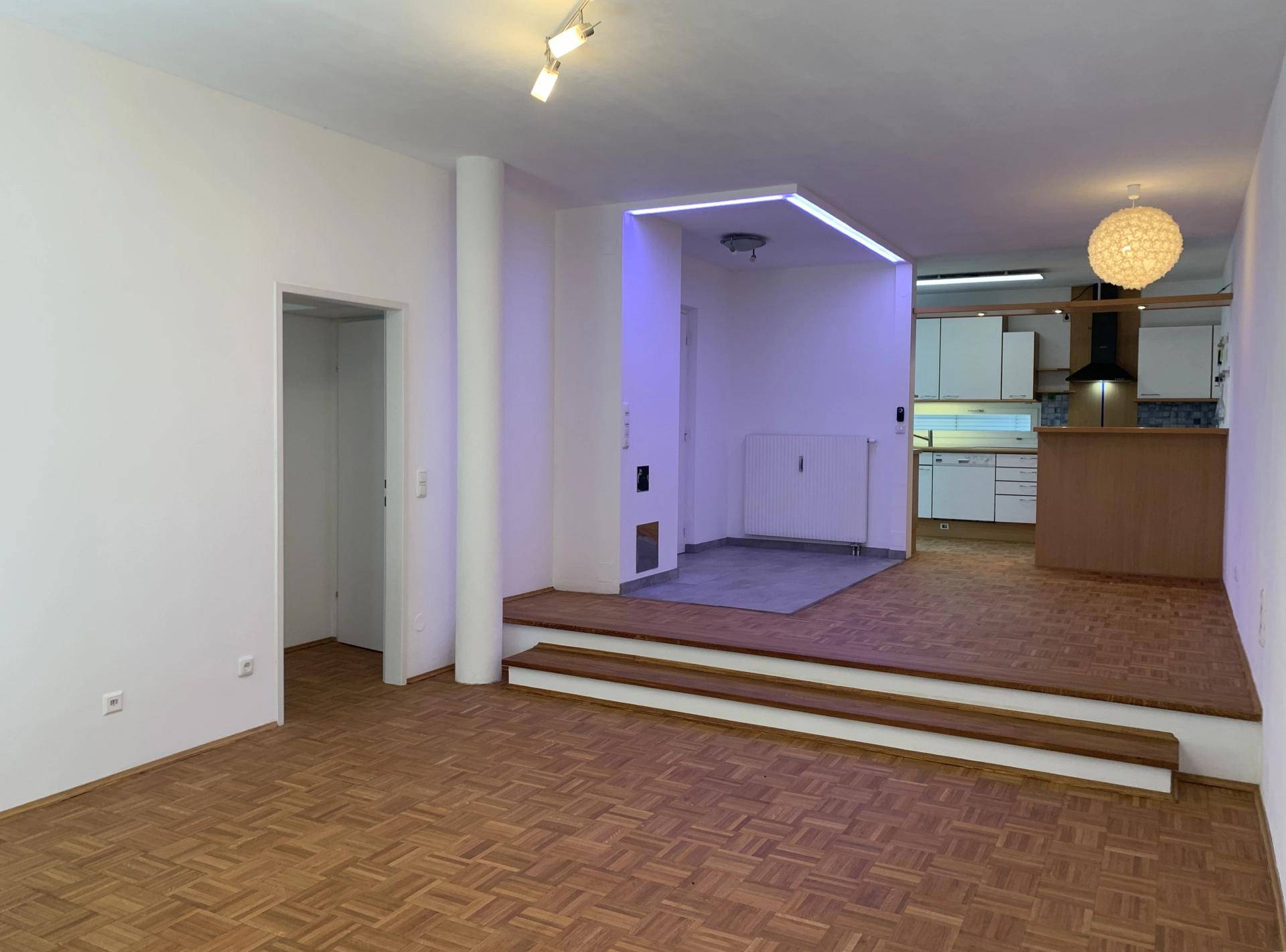 Offener Wohnbereich mit Parkettboden und einer erhöhten Fläche, die zur Küche führt.