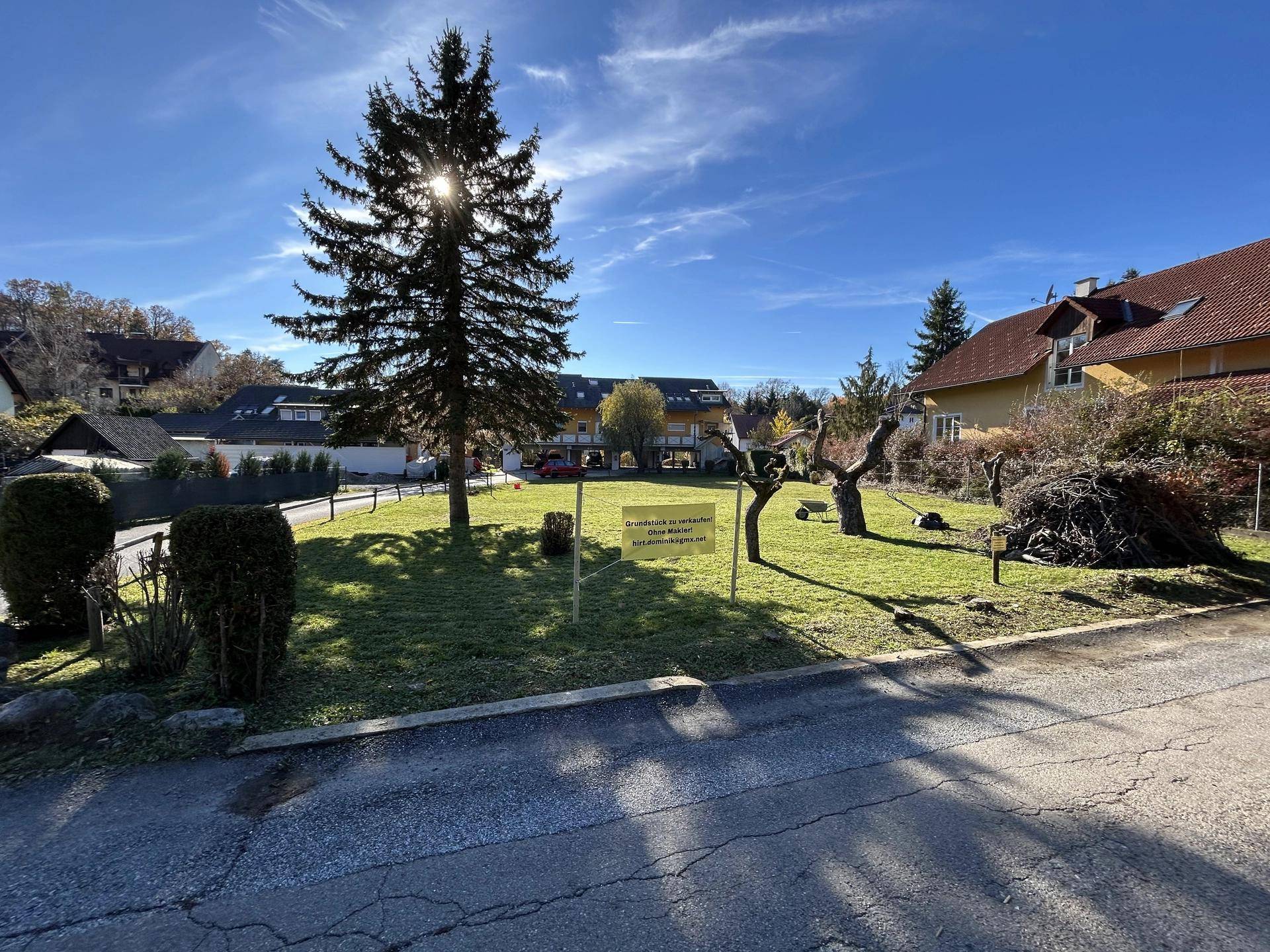 Helles Grundstück mit Verkaufsanzeige, umgeben von Wohnhäusern und Bäumen, unter blauem Himmel.