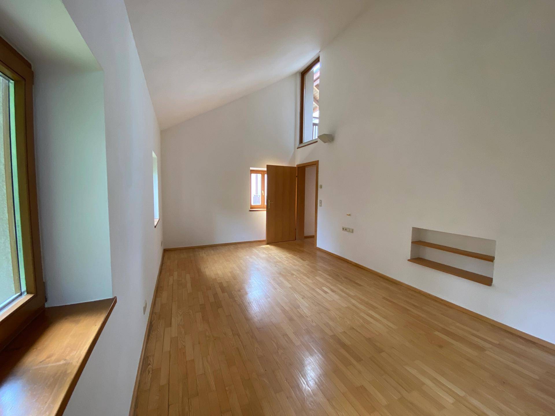 Heller Raum mit Holzboden, schrägen Decken und mehreren Fenstern.