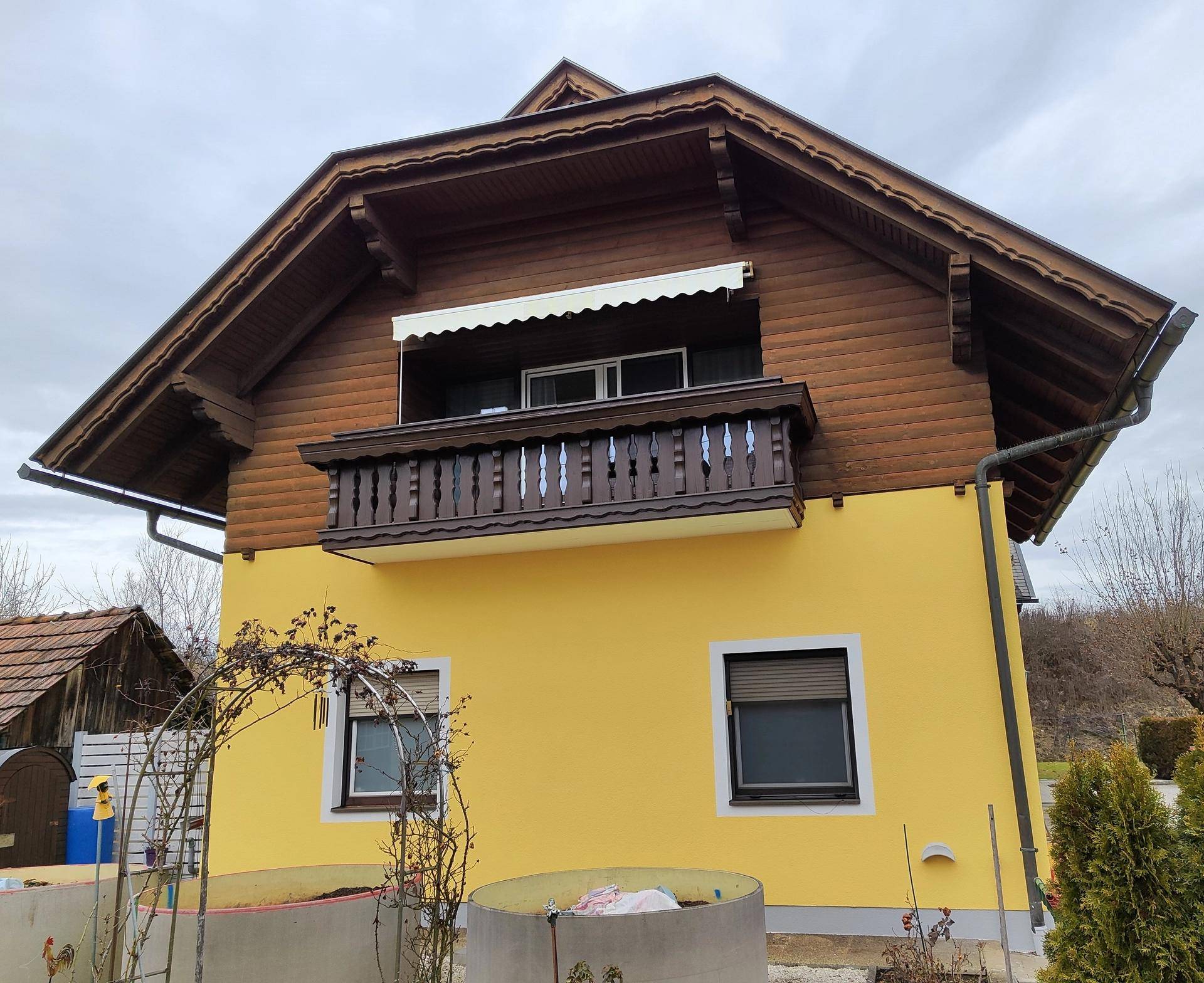 Seitenansicht des Gebäudes mit gelber Fassade, Holzbalkon und einer ausgefahrenen Markise.
