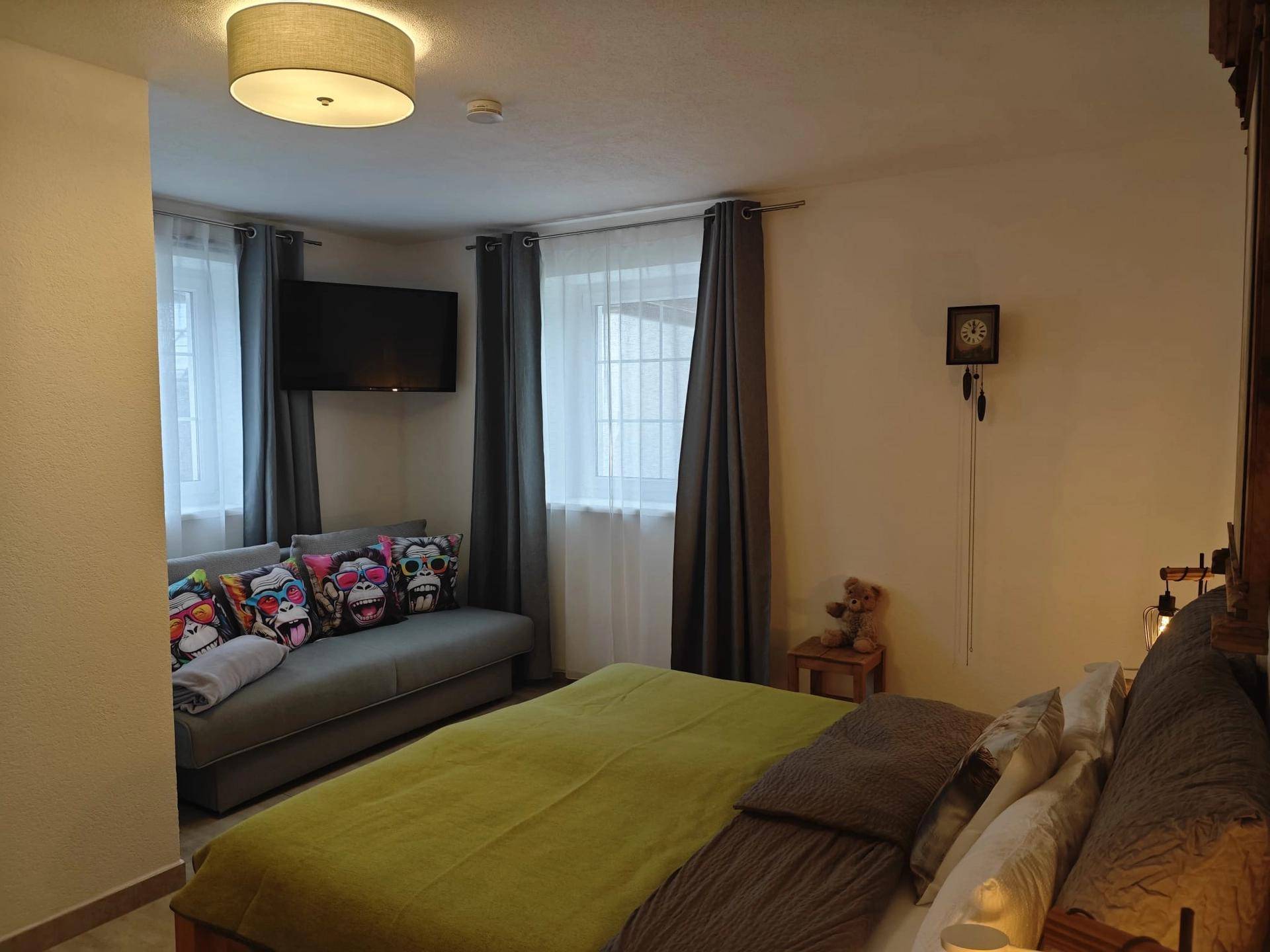 Gemütliches Schlafzimmer mit großem Bett, Sofa, zwei Fenstern und einem Wandfernseher.