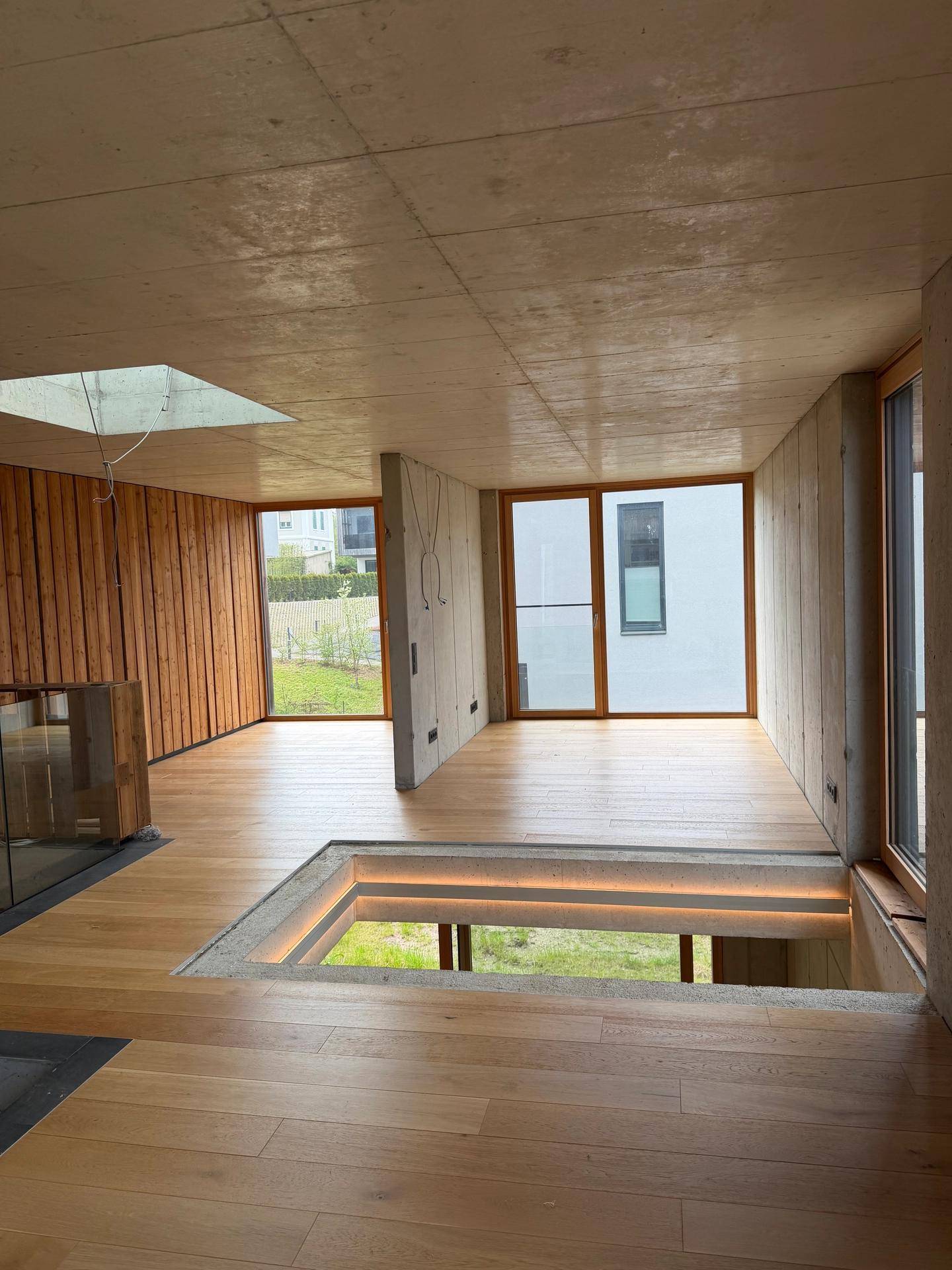 Heller offener Raum im Obergeschoss mit Holzboden, Sichtbetondecke und großen Fenstern mit Blick ins Grüne.